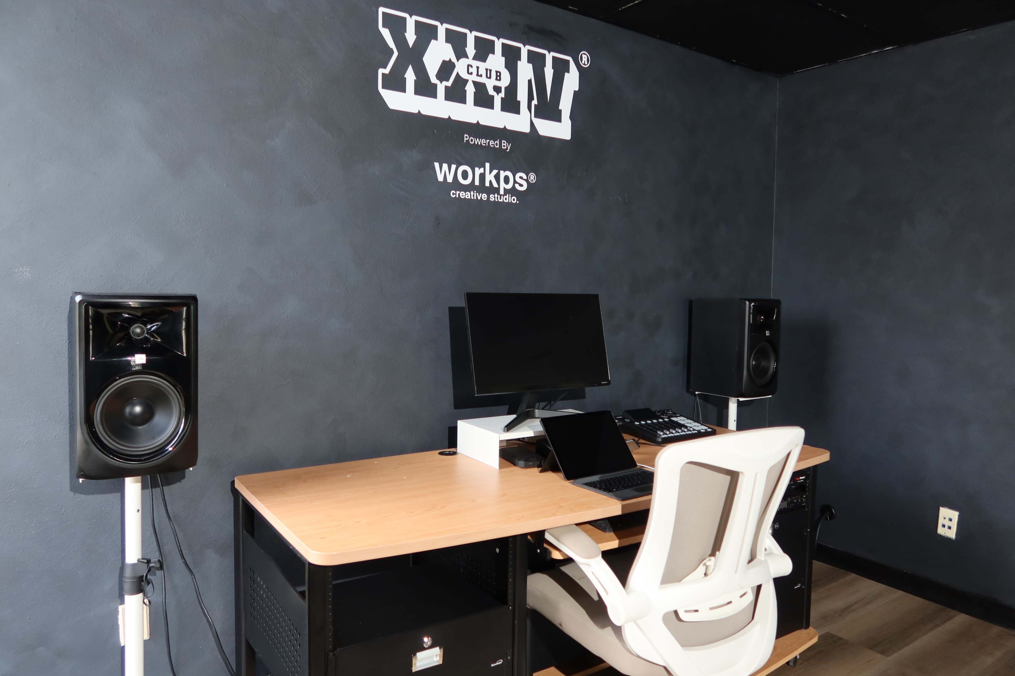 The image shows a minimalist audio studio with a desk, computer, and speakers on either side against a dark wall.