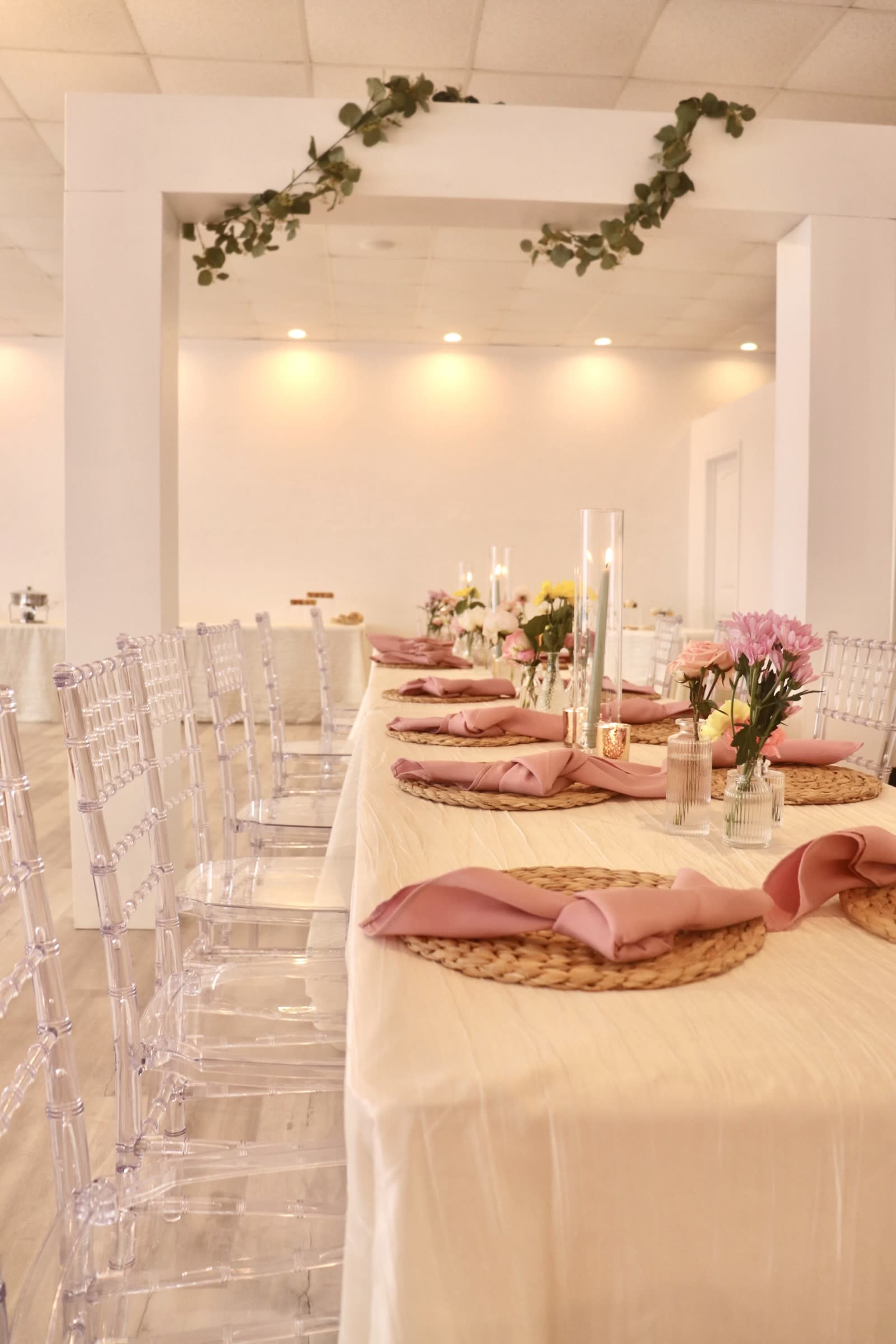 A long dining table is set with pink napkins, woven placemats, and clear chairs, adorned with floral centerpieces and candles.