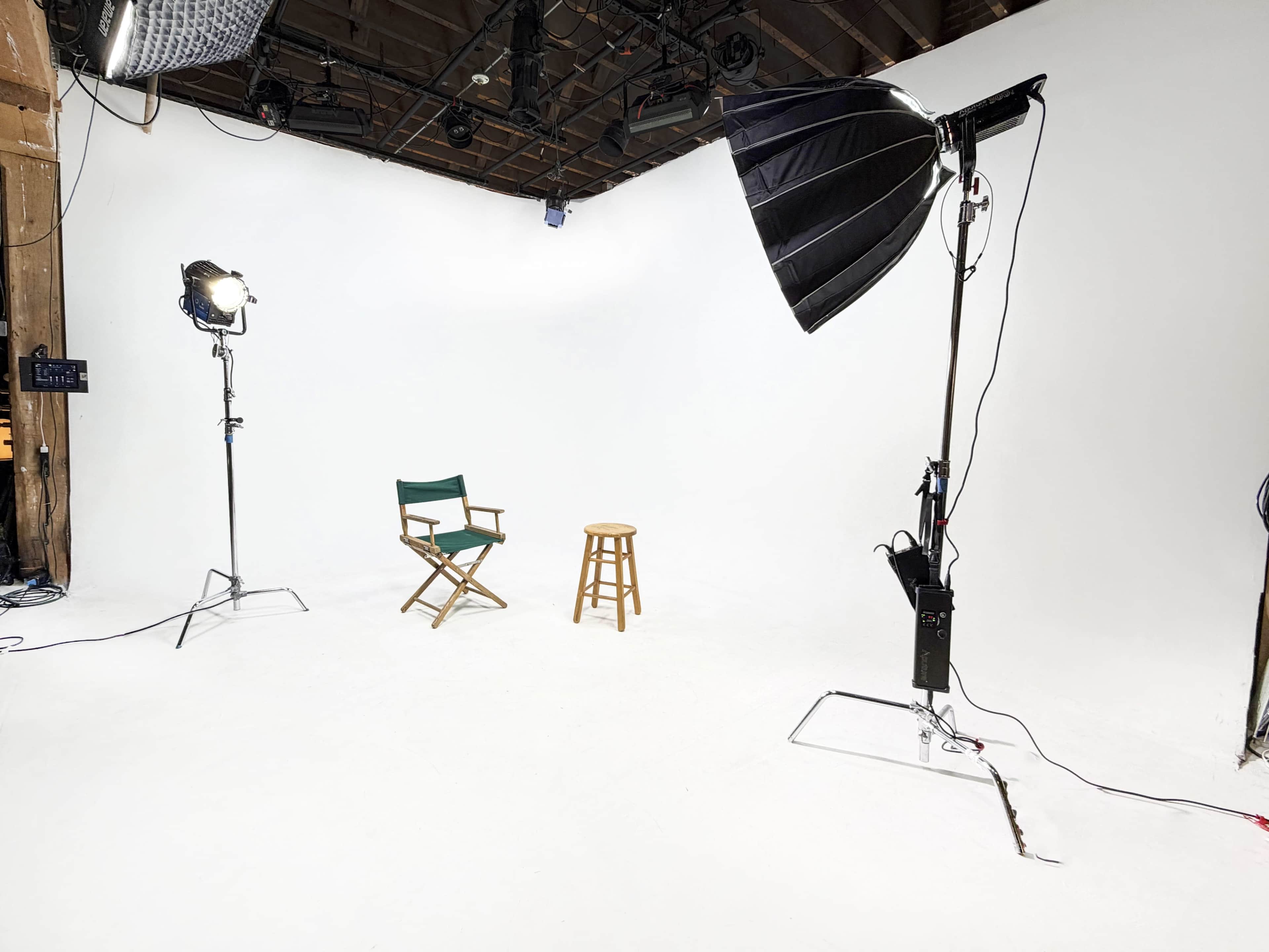 The image shows a photography studio setup featuring a white backdrop, a wooden stool, a director's chair, and various lighting equipment.