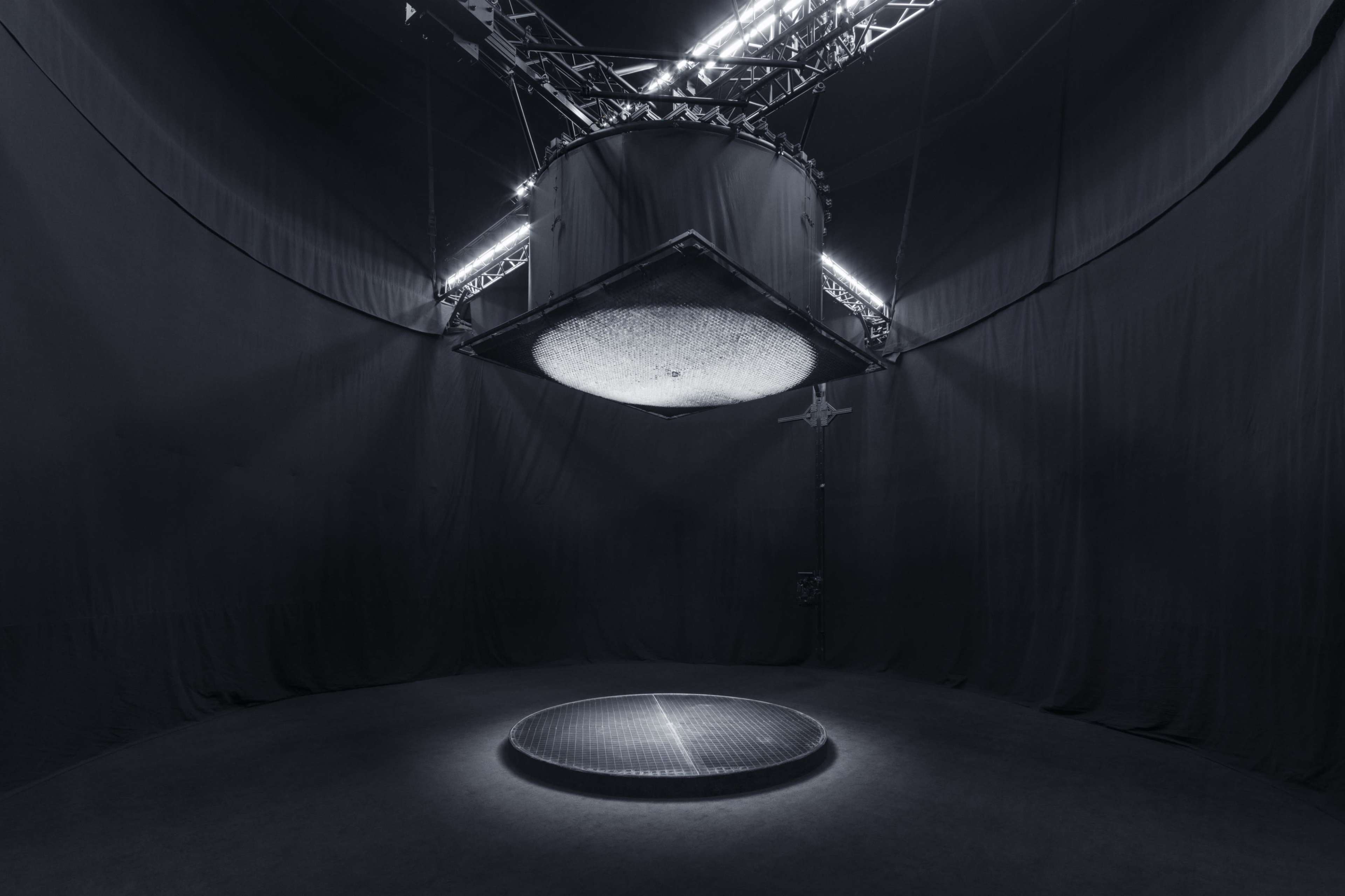 A dimly lit, circular platform positioned beneath a large, suspended light fixture inside a darkened space.