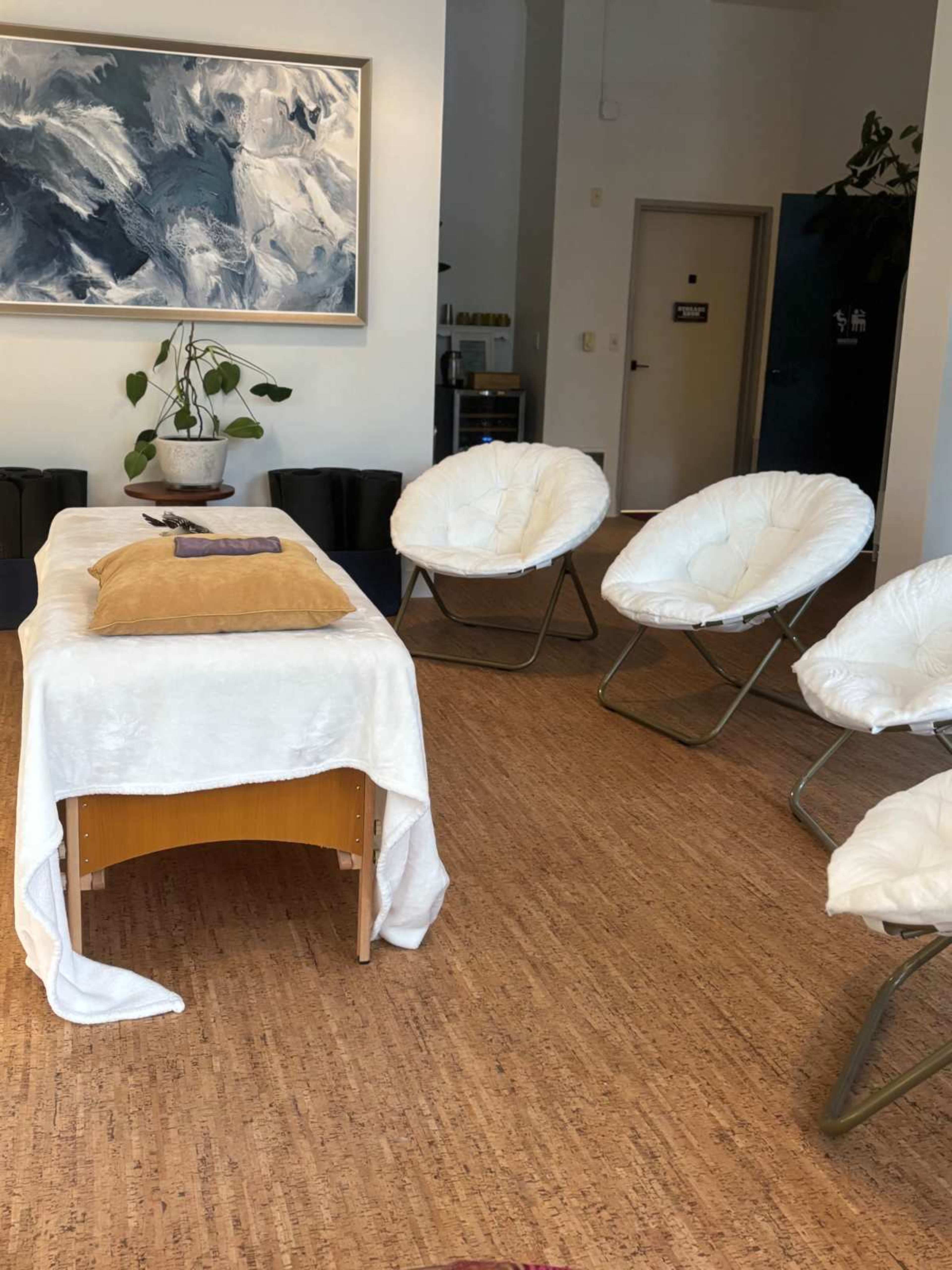 The image shows a serene room with a massage table covered in a white sheet and several white, fluffy chairs arranged around it.