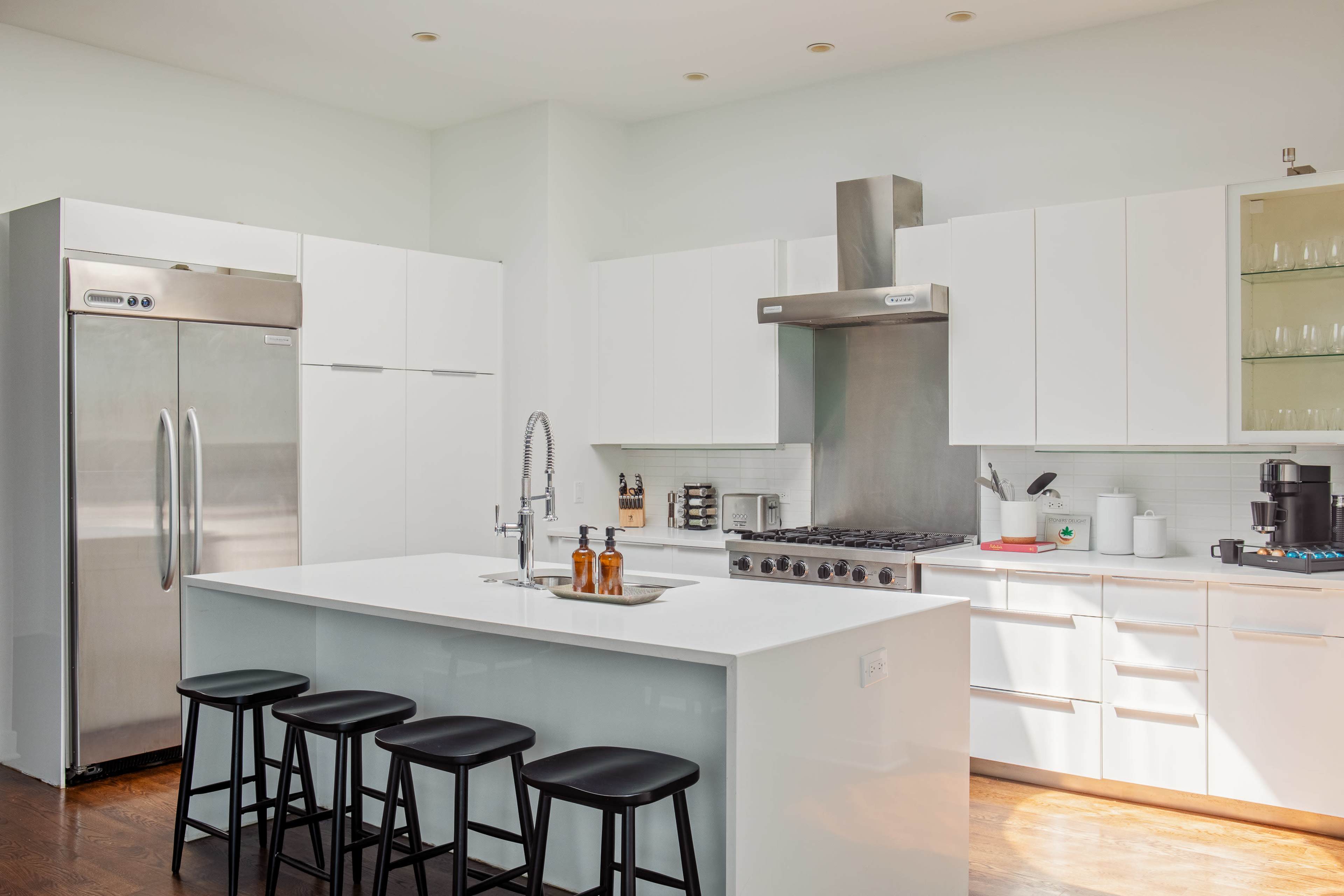 The kitchen features a modern design with a gray stainless steel range hood, white cabinets, a large island with four black stools, and stainless steel appliances.