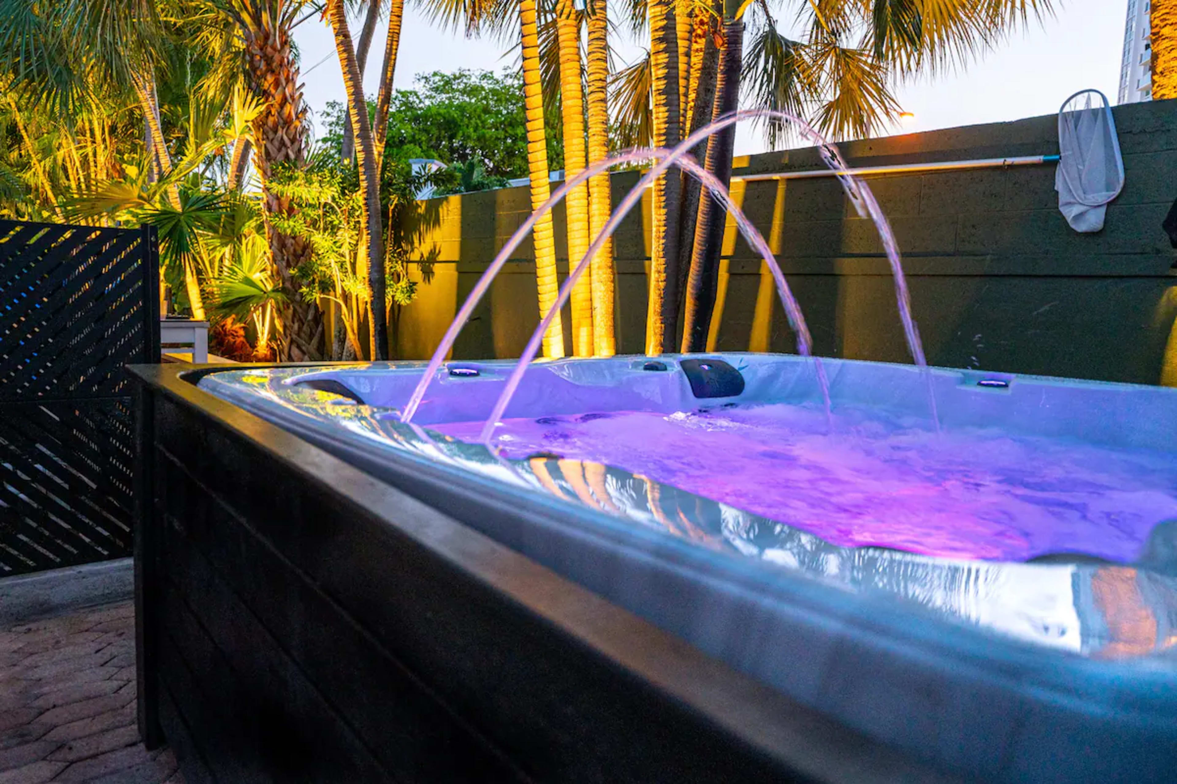 A spa tub with illuminated water jets is surrounded by palm trees and a wooden fence.