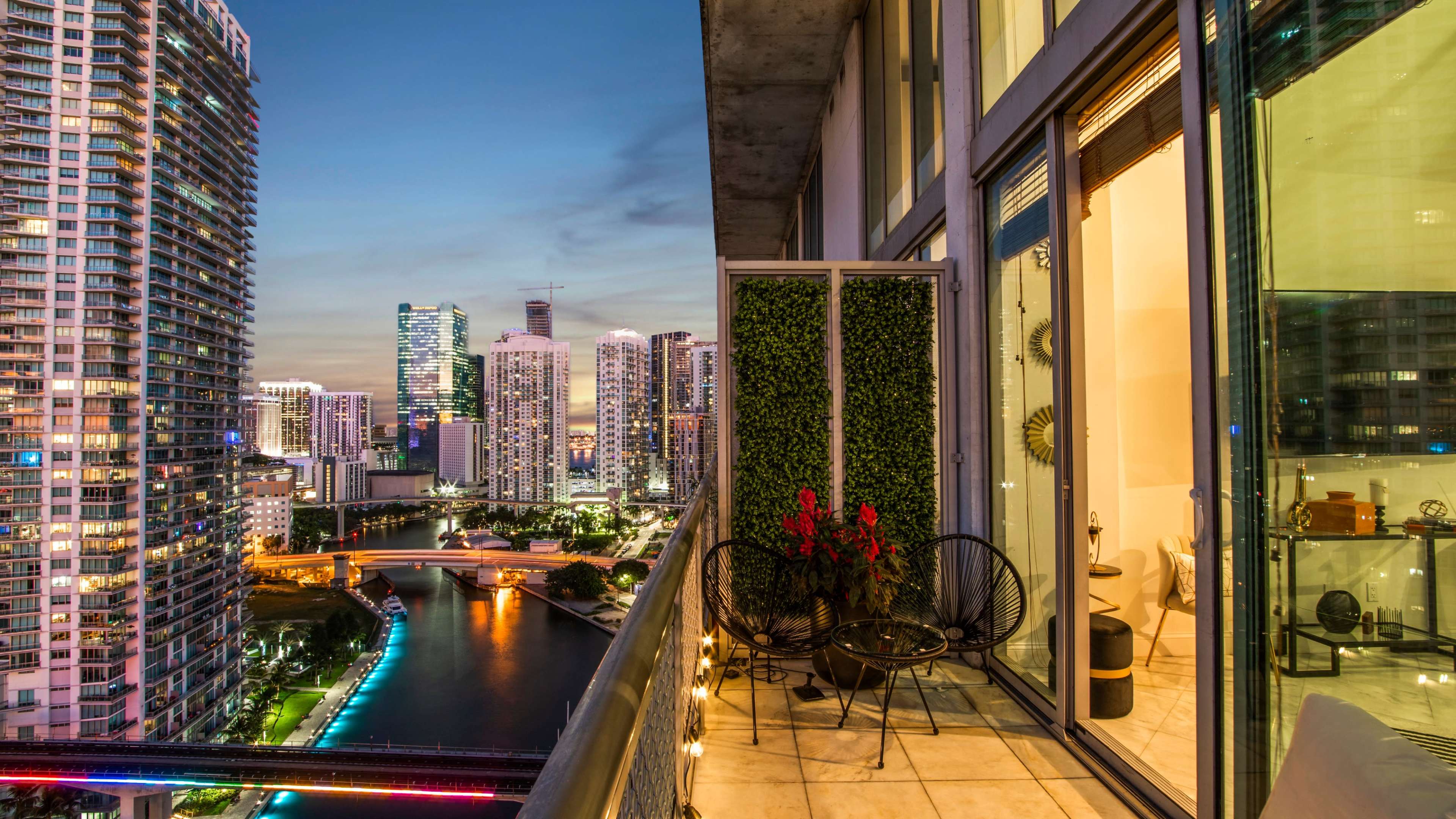 A balcony overlooks a city skyline at dusk, featuring tall buildings and a river, with a small seating area adorned with plants and lights.