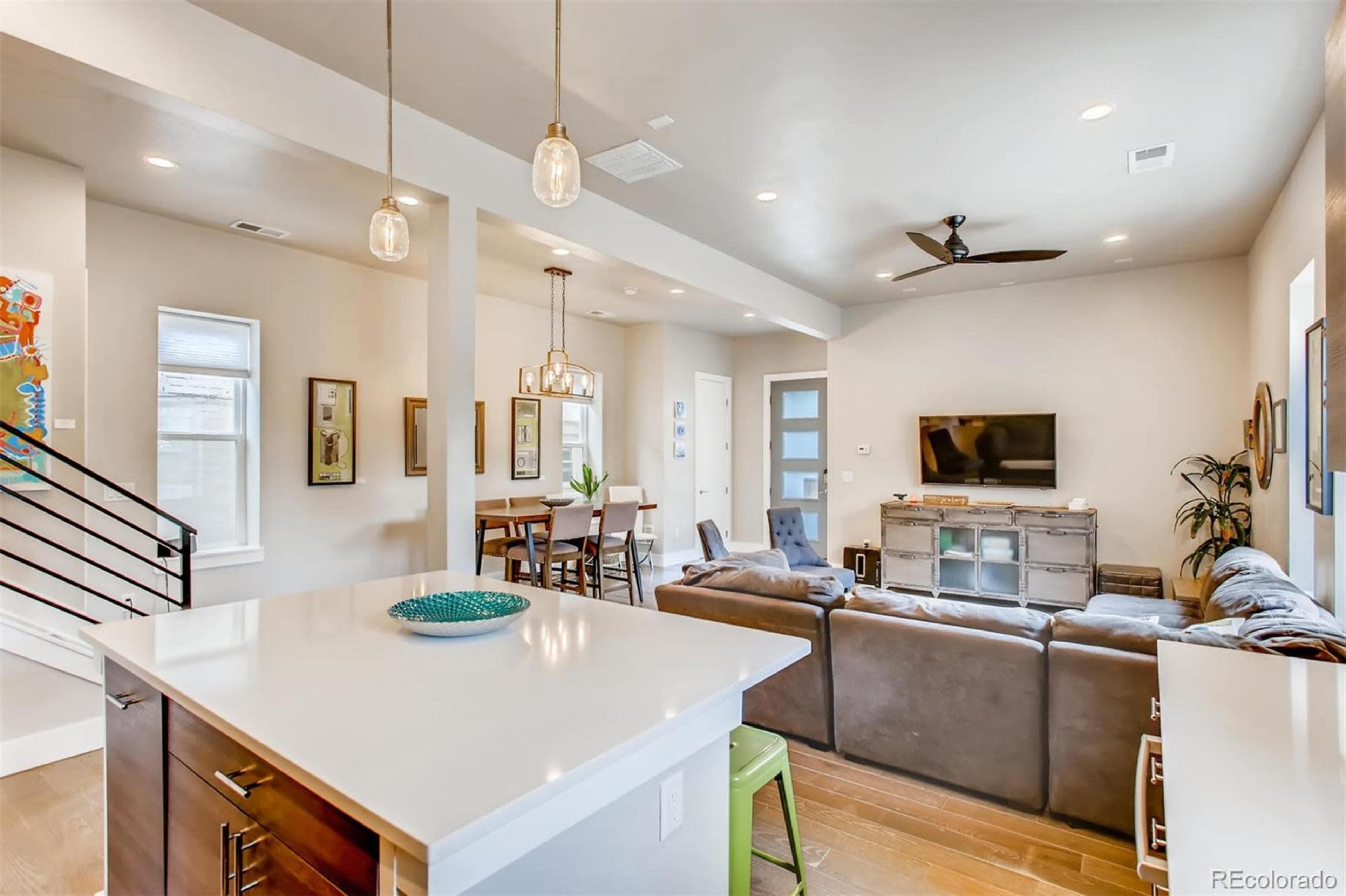 The image shows a modern living room and dining area featuring a sectional sofa, a wooden dining table, and large windows allowing natural light.