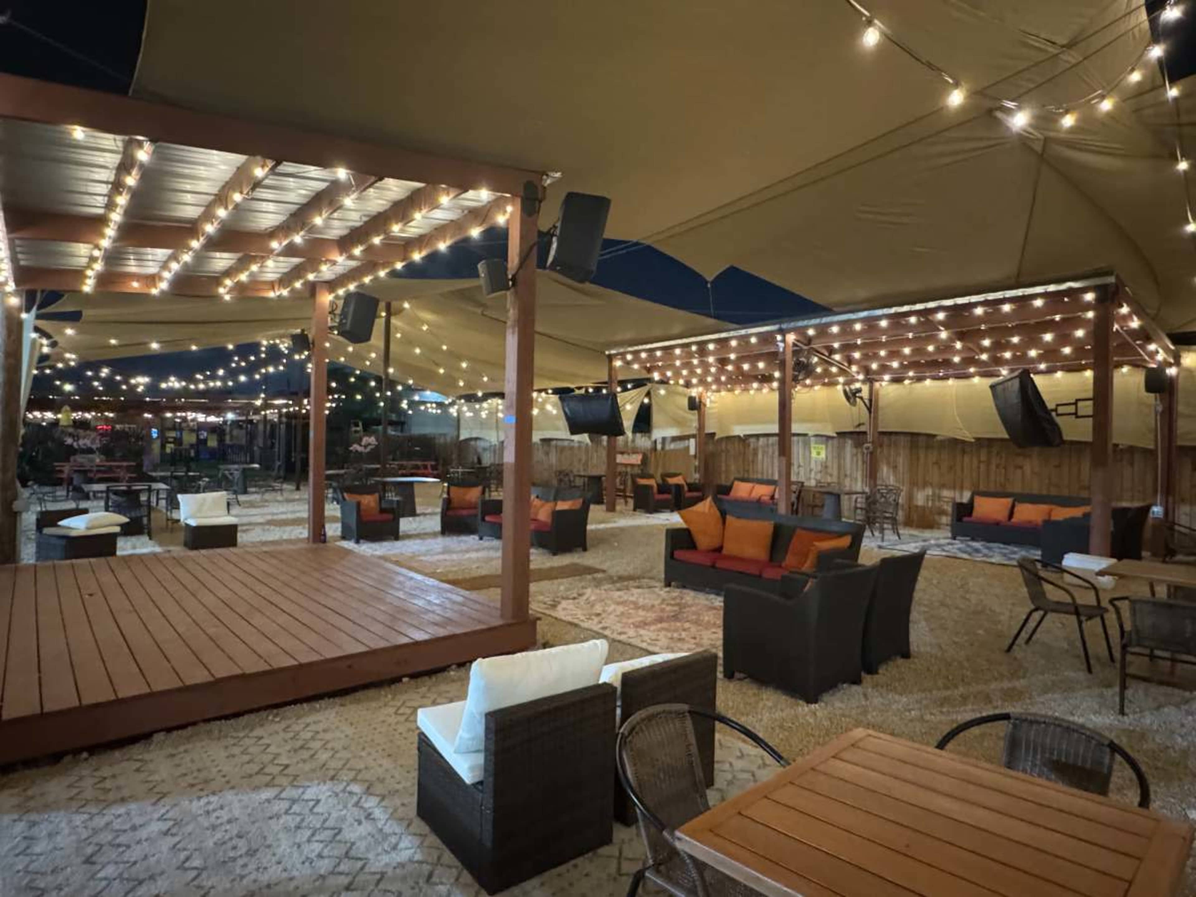 The image shows an outdoor seating area with wooden platforms, string lights overhead, and various furniture arranged on a gravel surface.