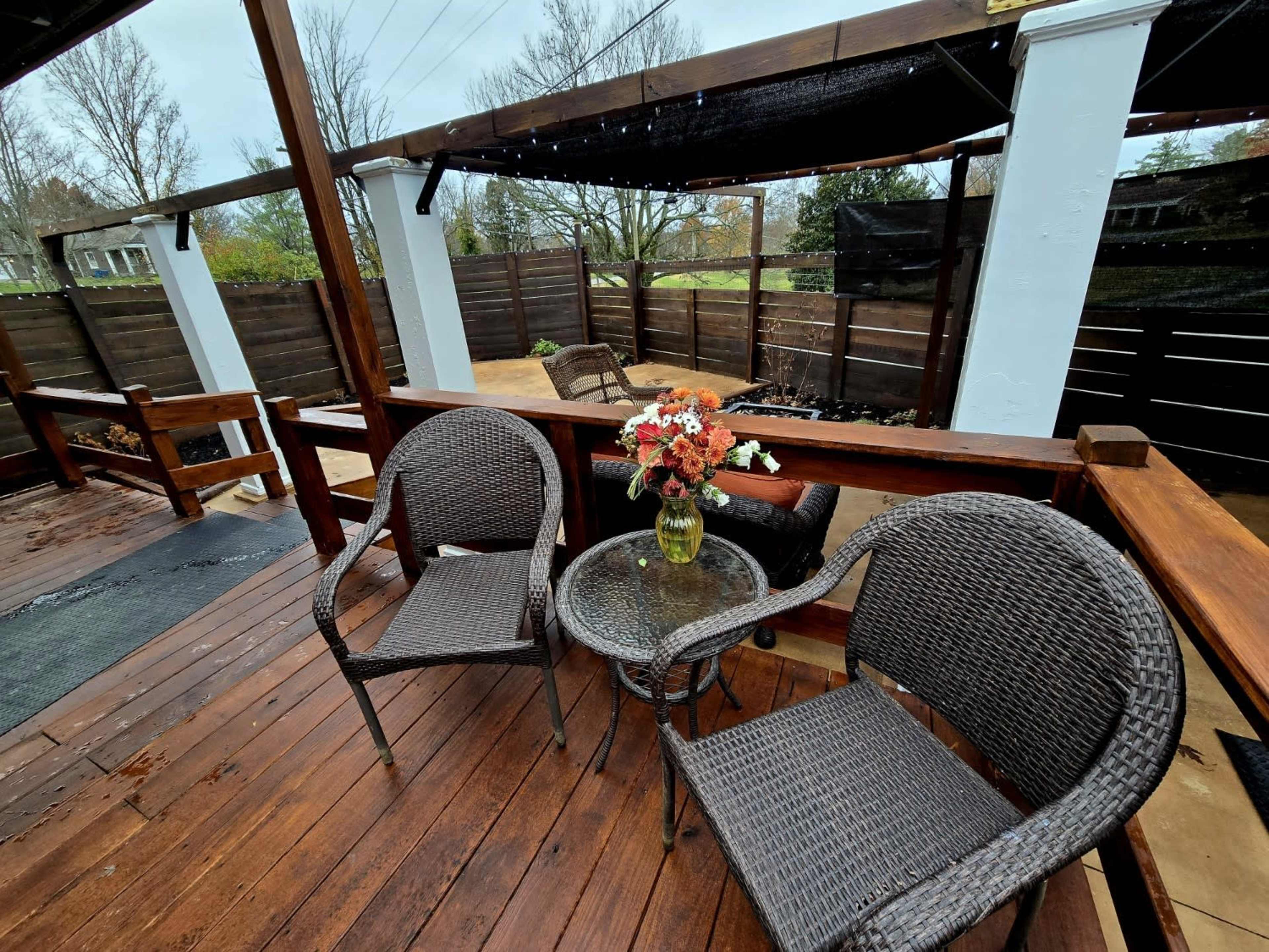 A wooden patio features two woven chairs and a glass table, adorned with a vase of flowers, surrounded by a fenced outdoor area.