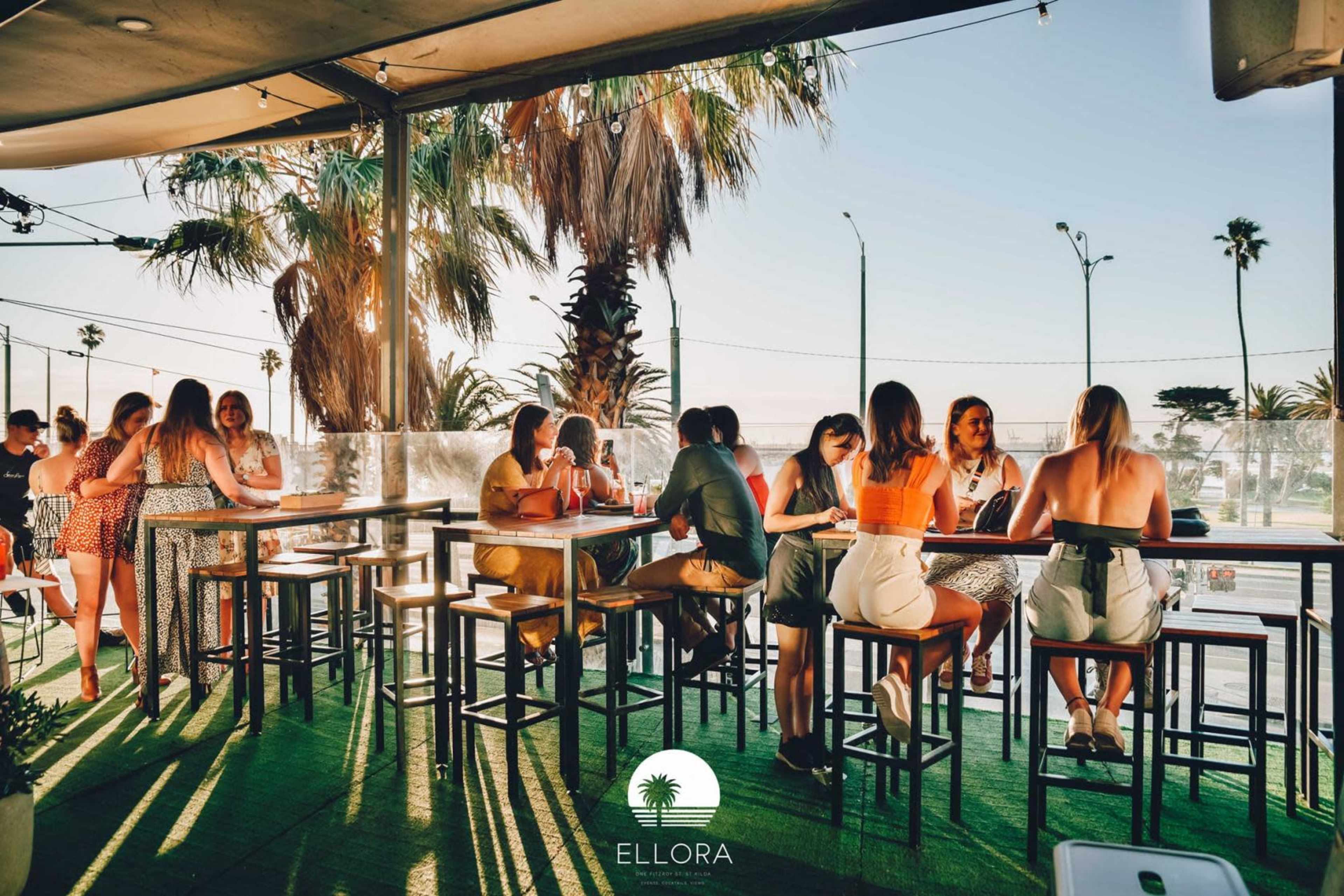 A vibrant outdoor dining area features a group of people gathered around tall tables, surrounded by palm trees and a sunset backdrop.