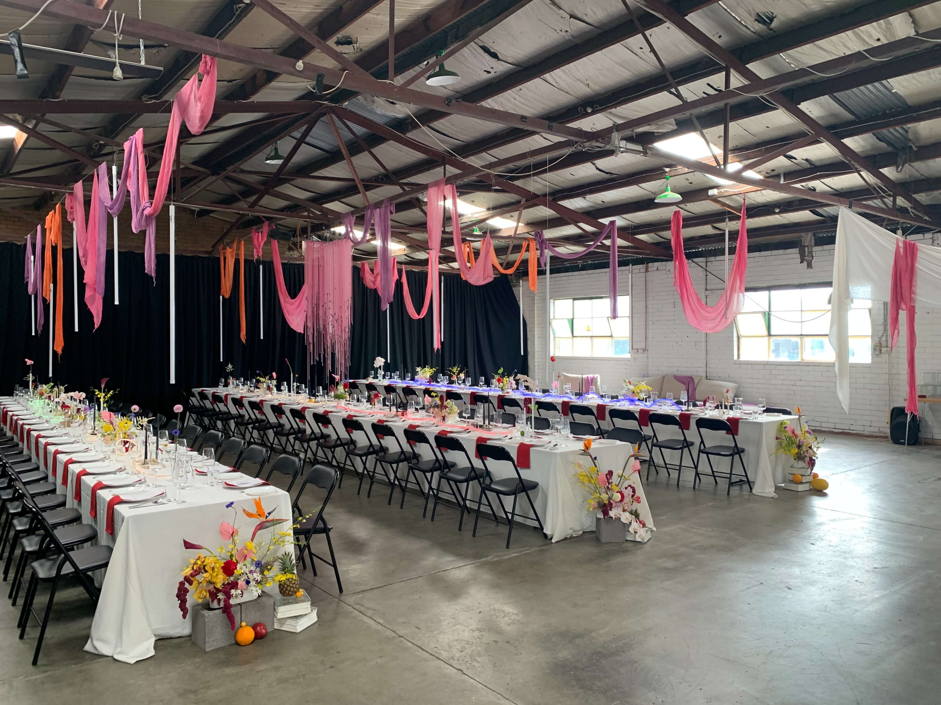 A spacious indoor event venue features long tables set for a formal gathering, adorned with flowers and draped fabric in shades of pink and orange.