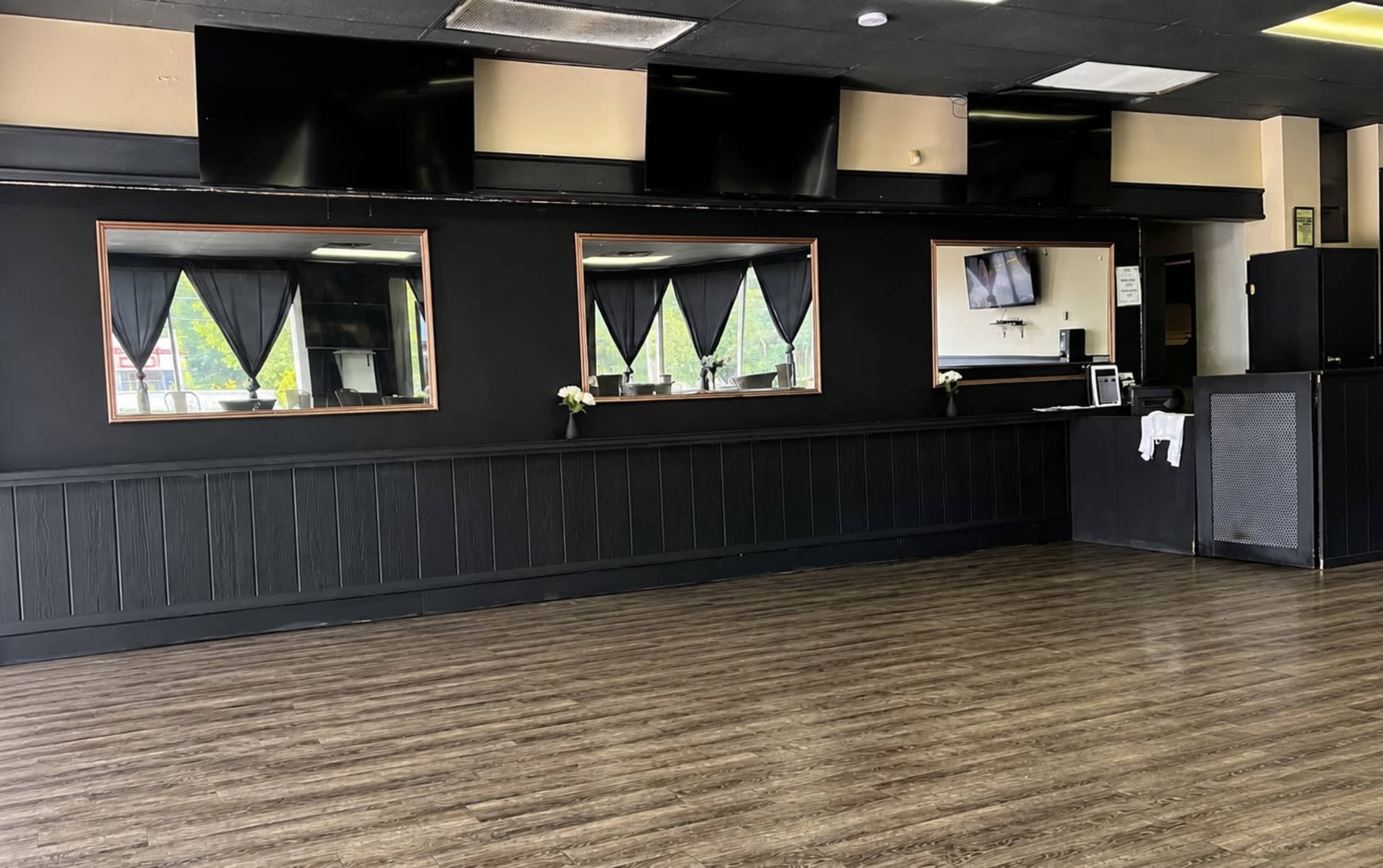 The image shows a sparsely furnished room with dark walls, wooden flooring, and two mirrors with black curtains above, alongside a service counter and a few monitors.