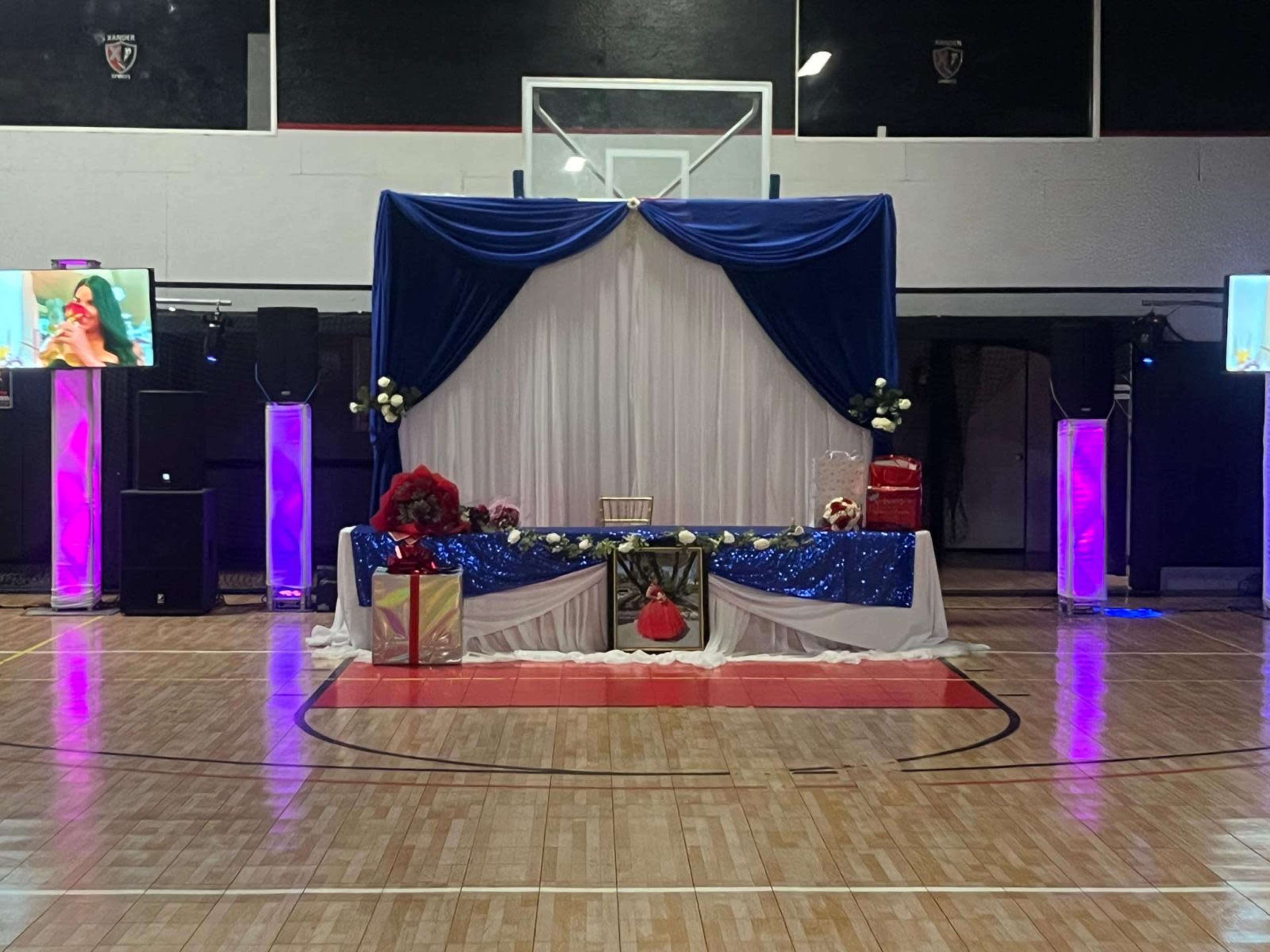 The image shows a decorated stage with a blue and white backdrop, floral arrangements, and a table adorned with presents, set up in a gymnasium.