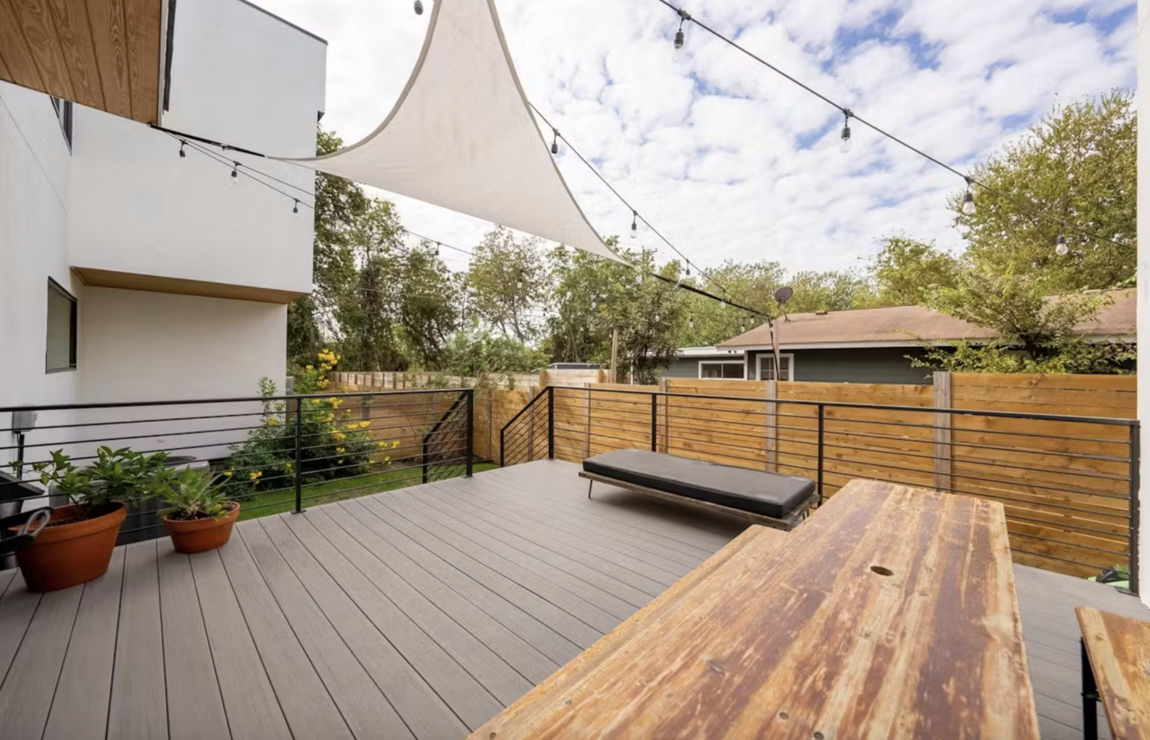 The image shows a spacious outdoor deck with a wooden table, a lounge chair, potted plants, and a large shade sail, surrounded by fenced greenery and a residential area.