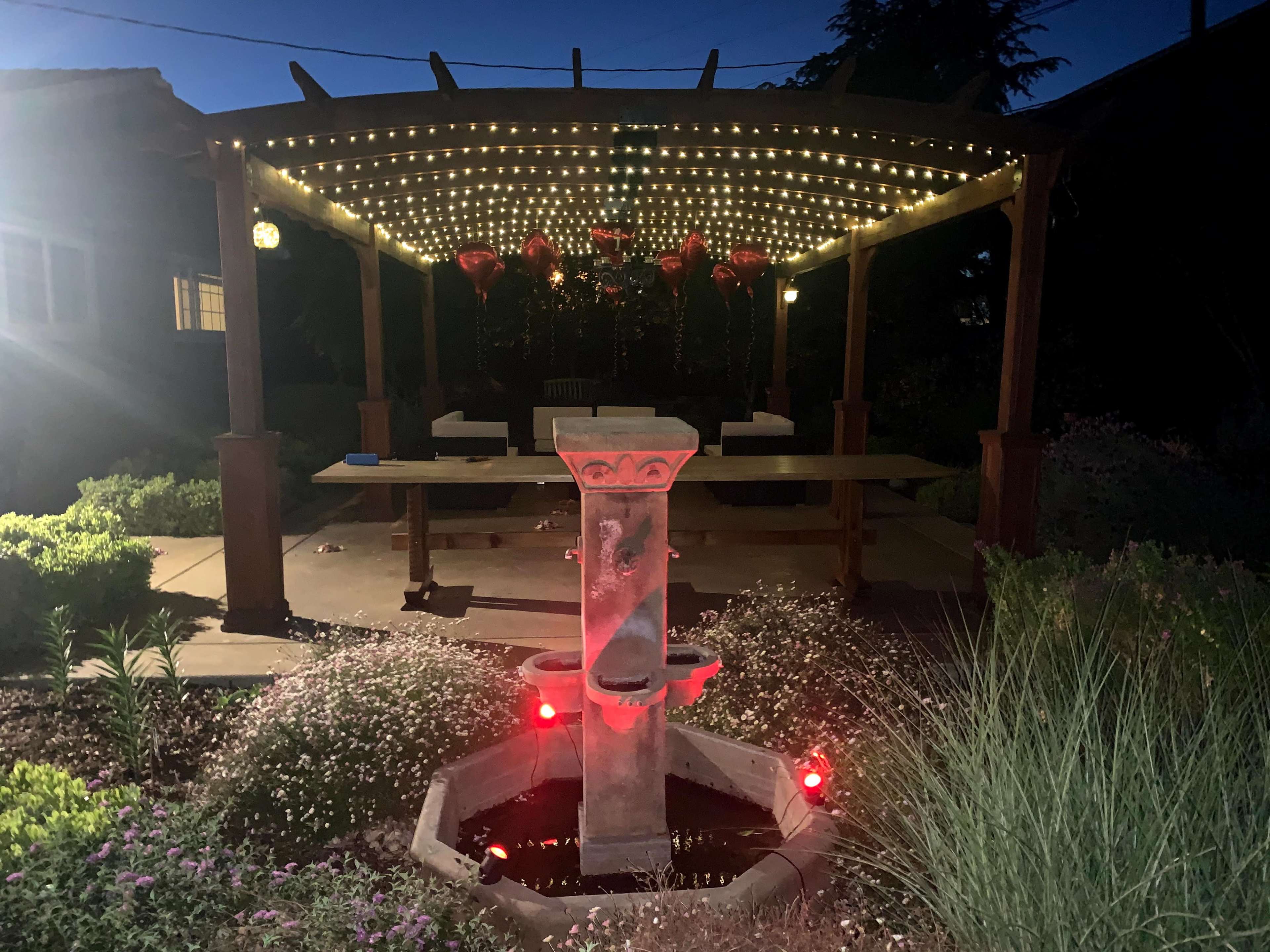 A wooden pergola with string lights and red decorations houses a stone fountain surrounded by landscaped plants at night.