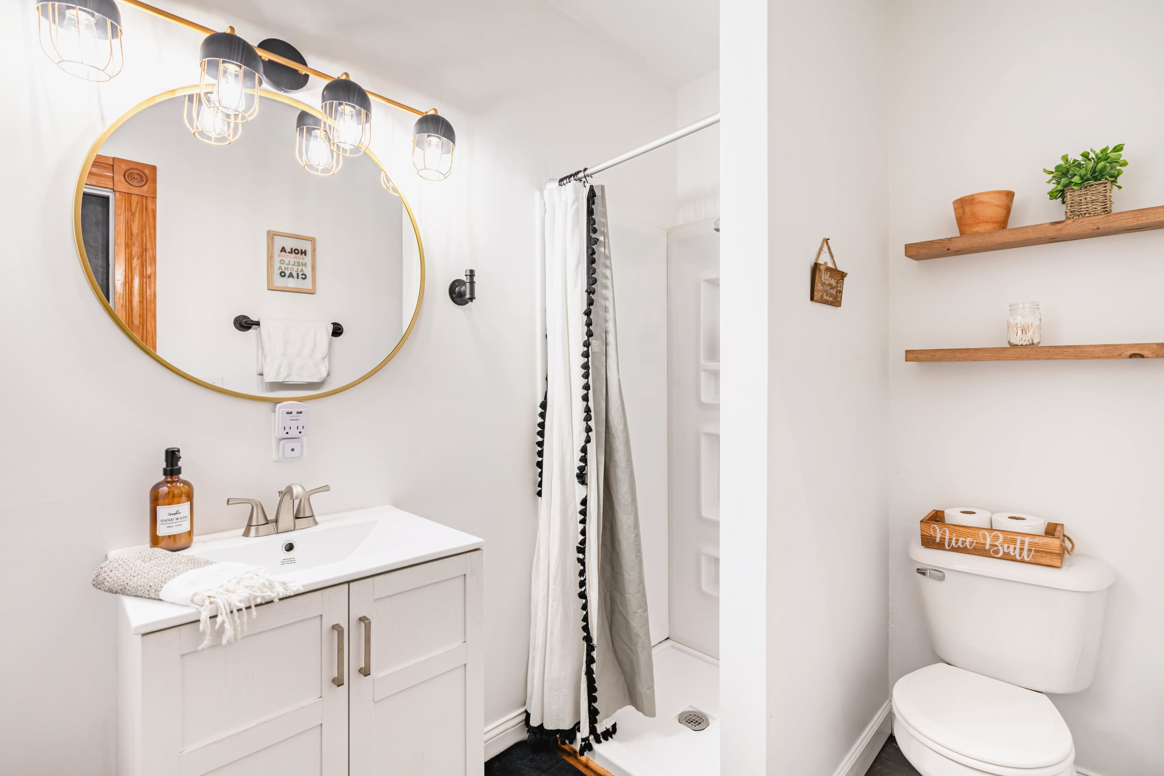 A modern bathroom featuring a shower, a round mirror with lights, a towel rack, and wooden shelves with decorative items.
