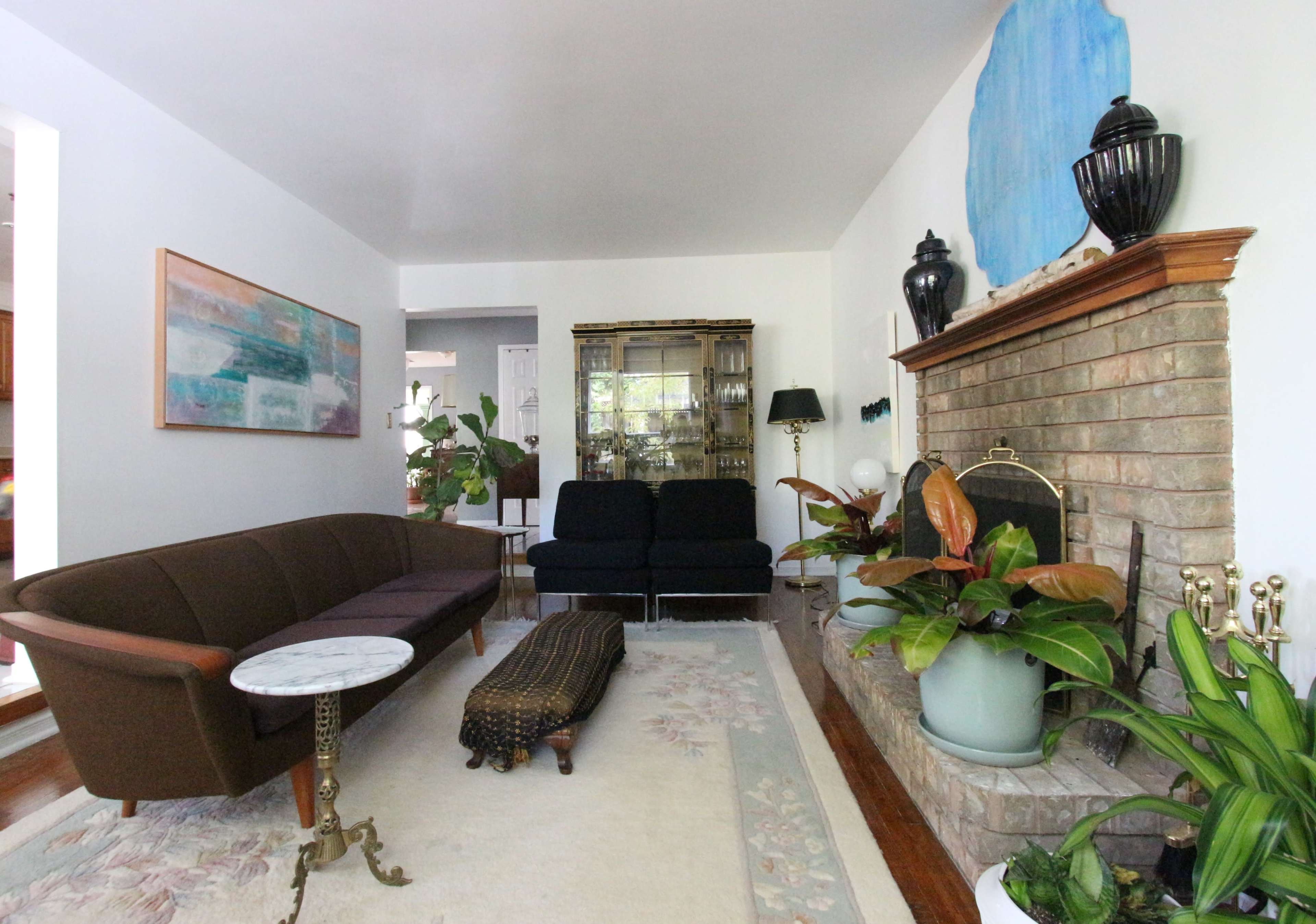 The living room features a brown couch, two black chairs, a stone fireplace with plants, and decorative artwork on the walls.