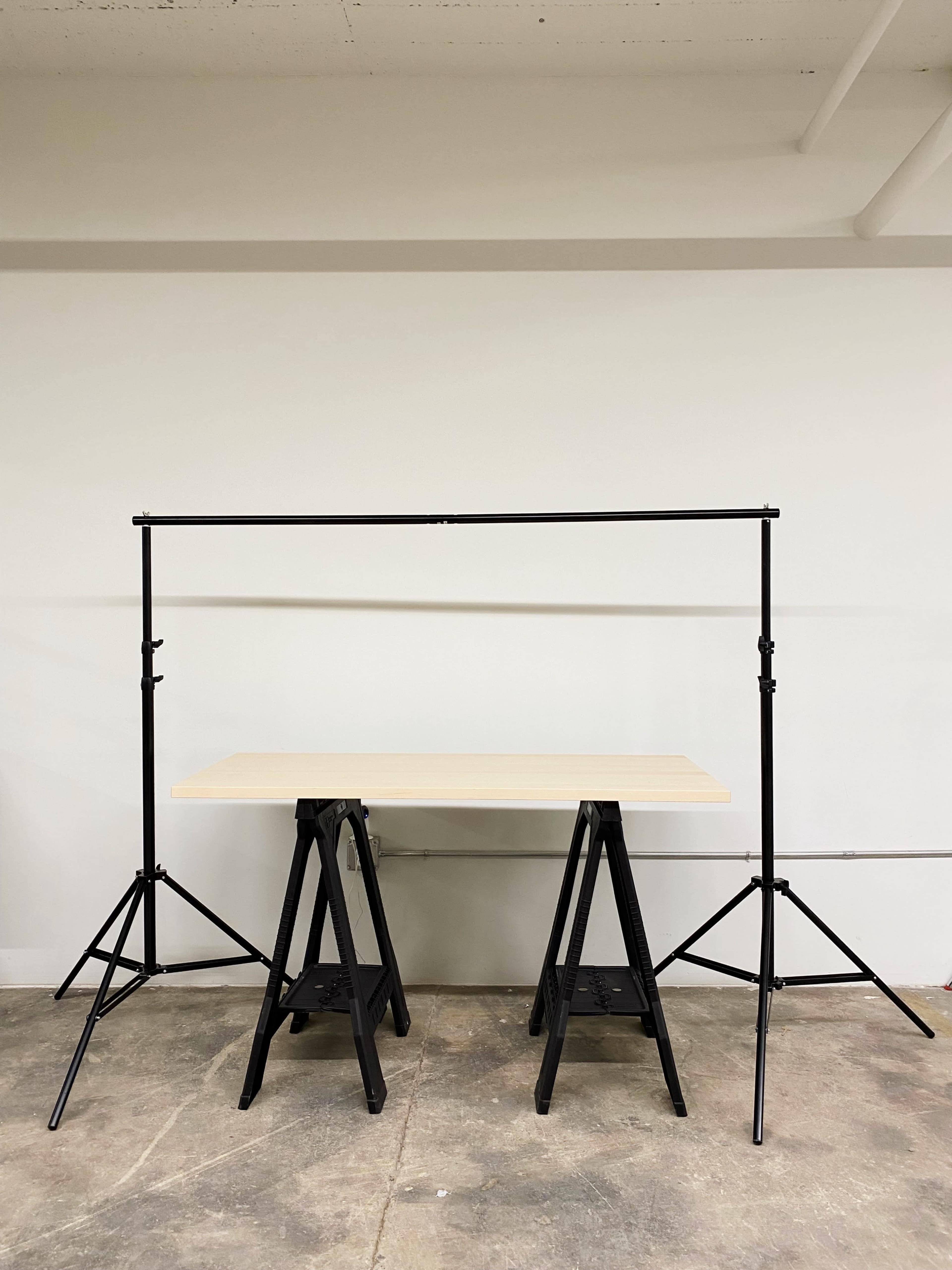 A wooden table supported by two black sawhorse legs, with a black backdrop support frame positioned behind it.