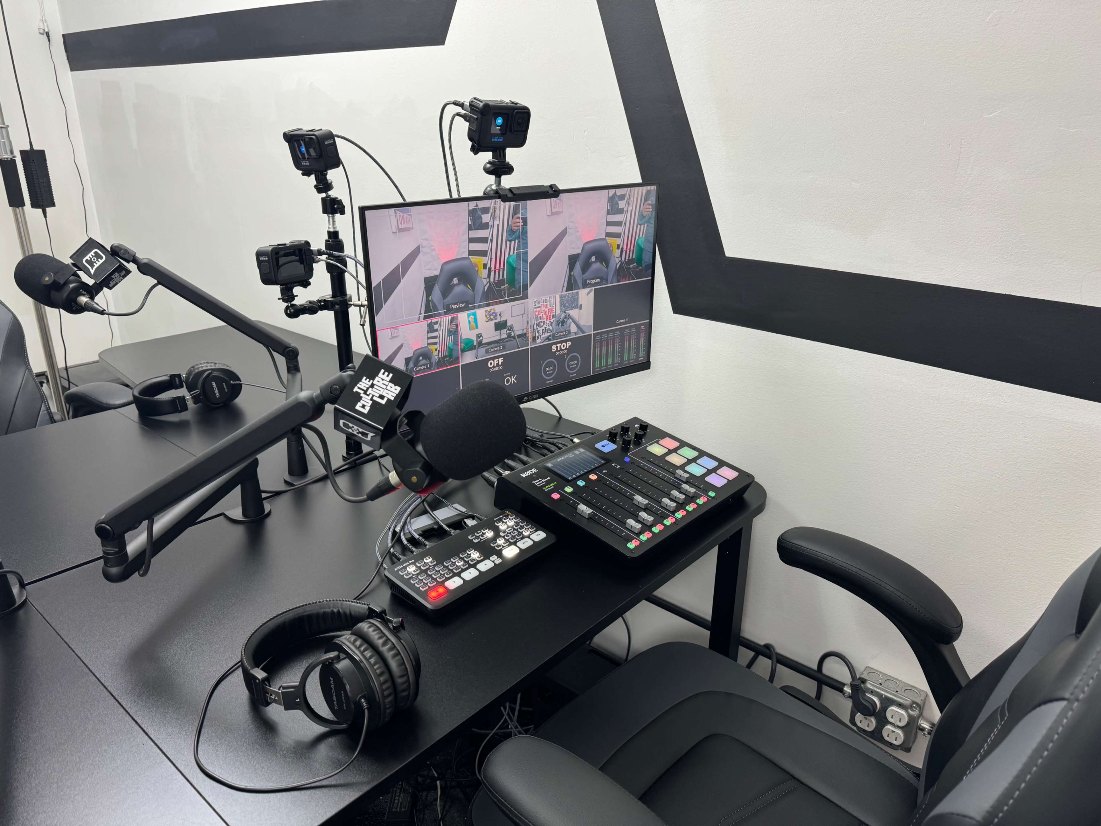A radio or podcasting studio setup, featuring a mixing console, microphones, headphones, and cameras on a black desk against a white wall with geometric designs.