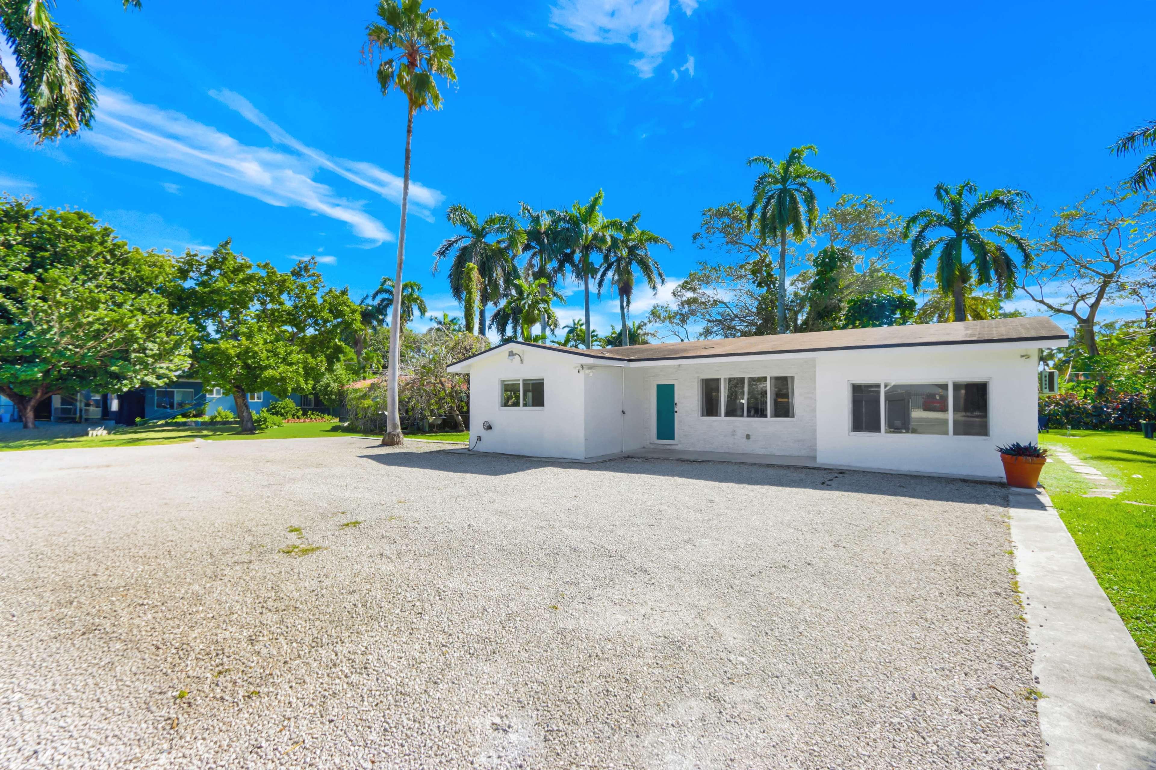 The image shows a single-story white house surrounded by palm trees and a gravel driveway in a sunny, open landscape.