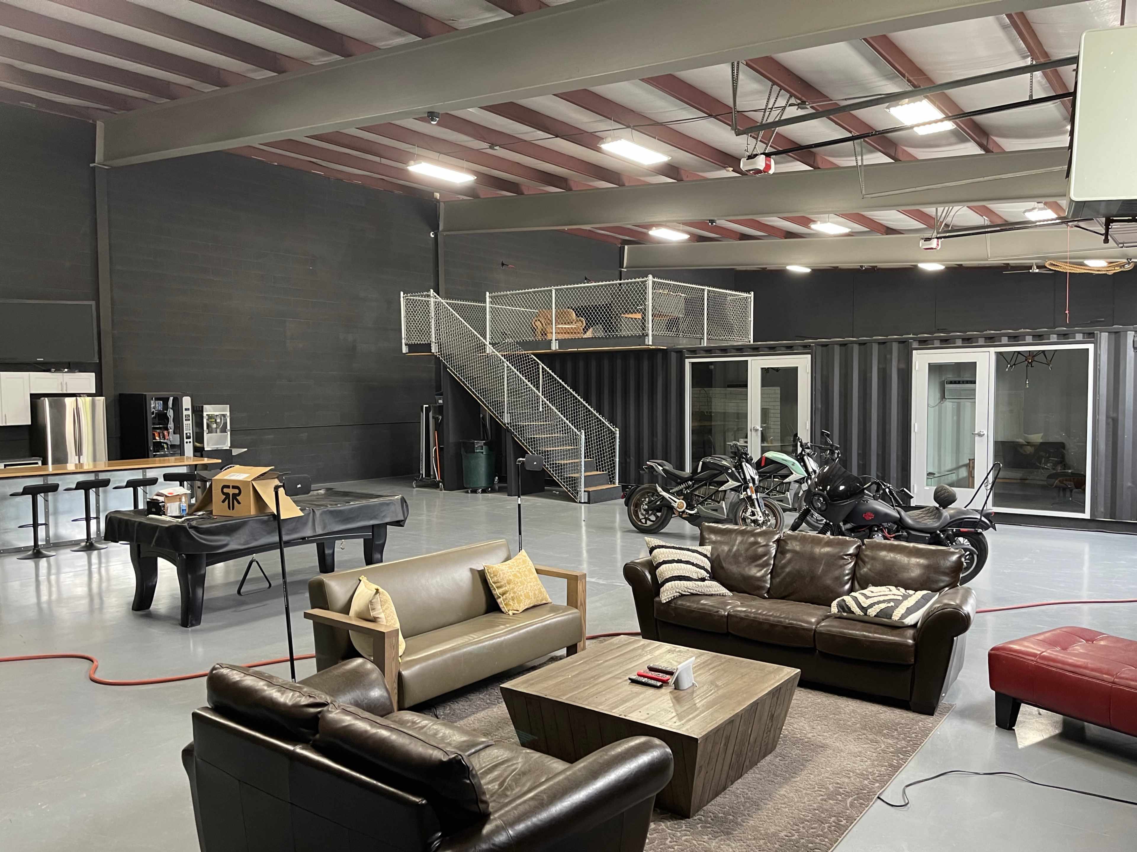 A spacious, industrial-style interior featuring a seating area with leather sofas, a coffee table, and a piano, along with a two-wheeled motorcycle parked nearby and a staircase leading to a mezzanine level.