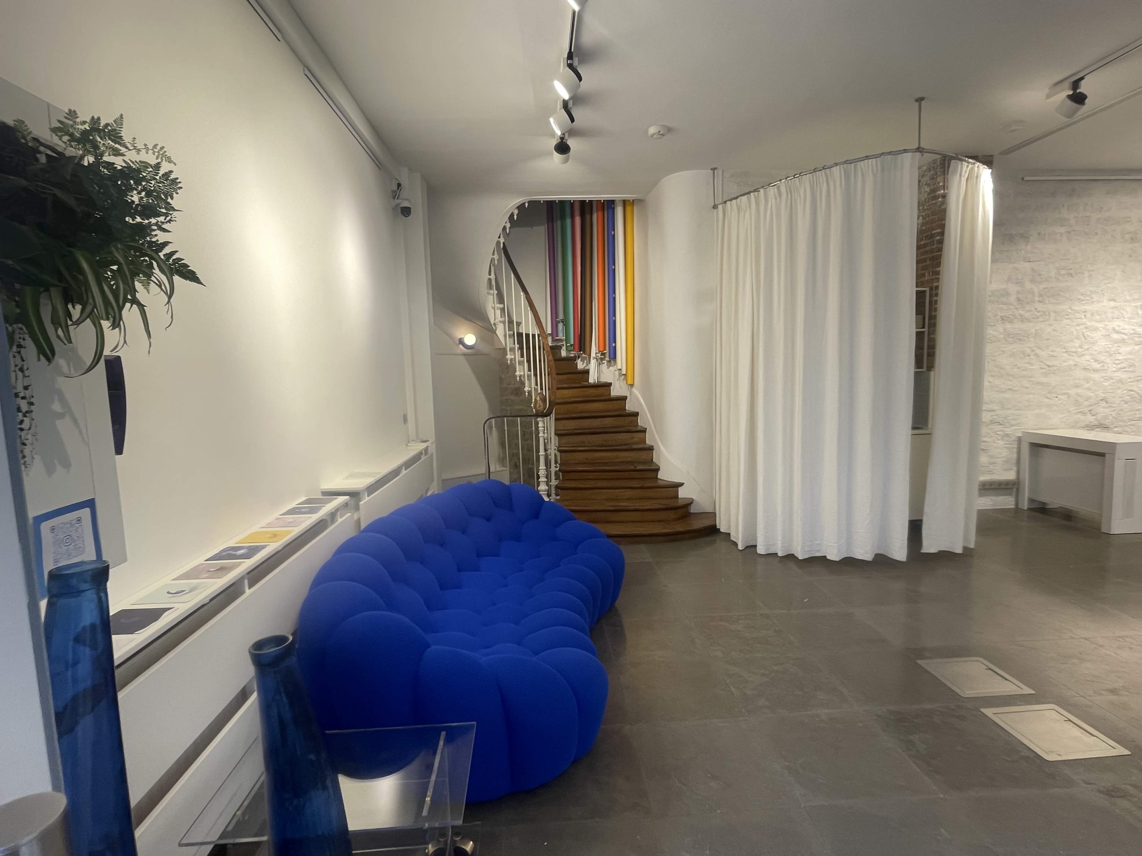 The image shows a spacious interior featuring a blue sofa, a staircase with colorful strips, and a curtain separating different areas.