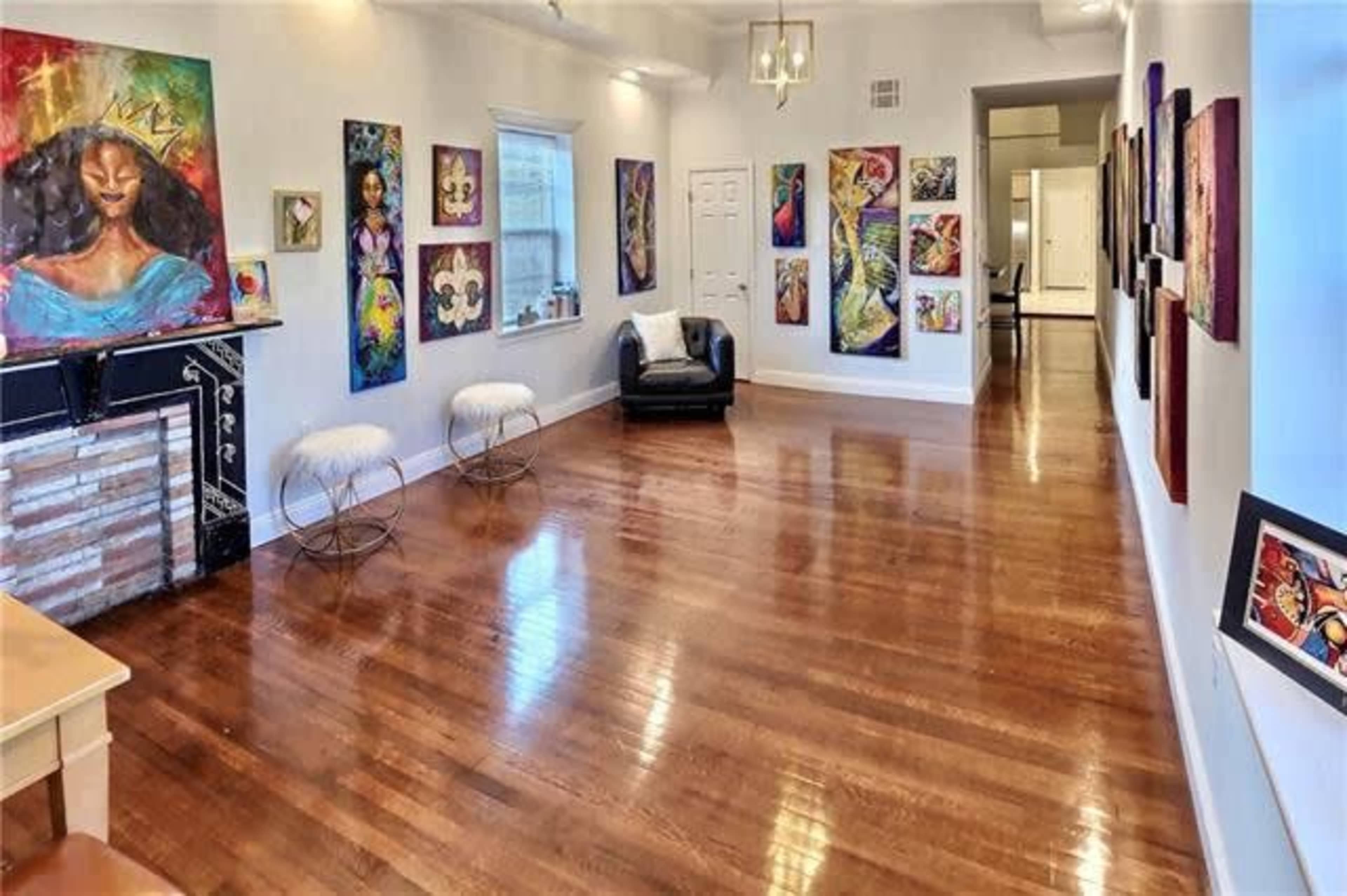 A brightly lit art gallery with wooden floors, featuring colorful paintings on the walls and a seating area with a black chair and two white stools.