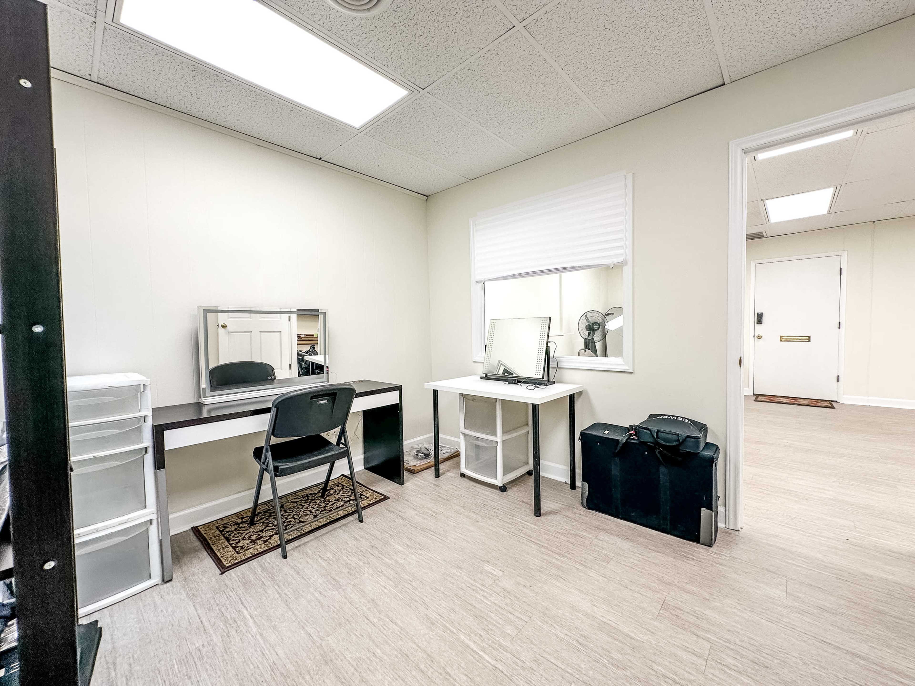 A simple office space with a mirror, two desks, a chair, and a window with blinds.