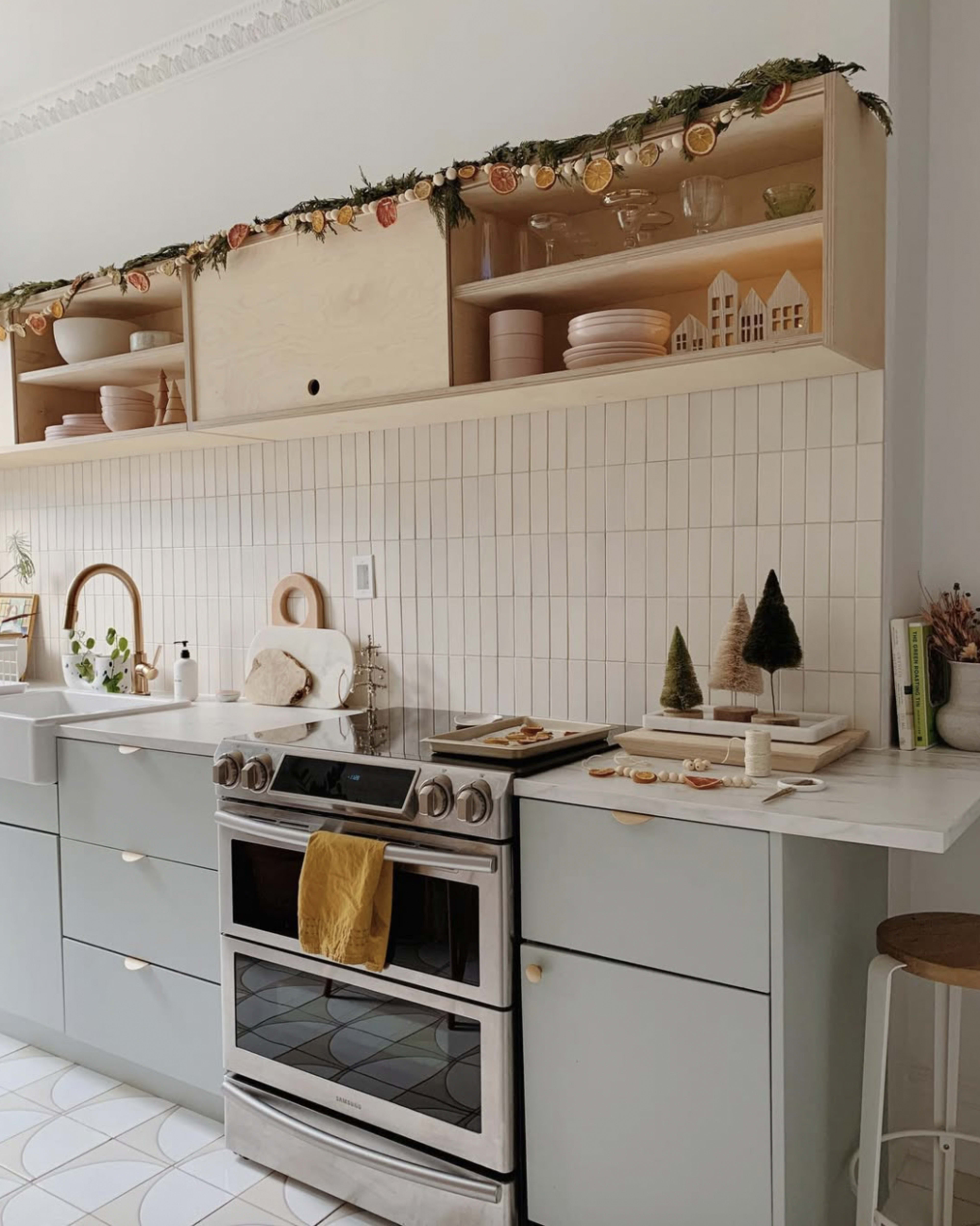 The kitchen features light-colored cabinets, a stainless steel oven, and open shelving adorned with decorative items and a garland.