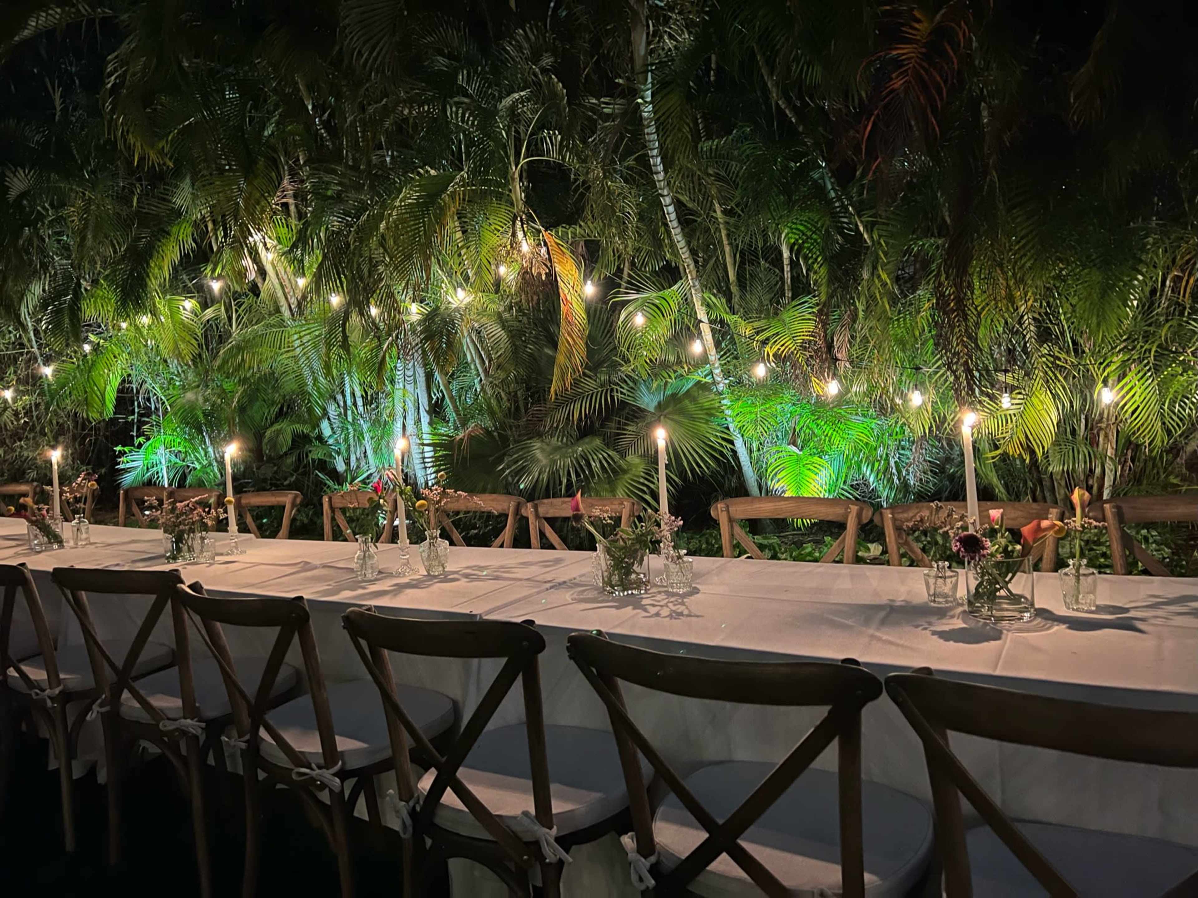 A long dining table is set outdoors, surrounded by tropical plants and illuminated by soft candlelight and string lights.