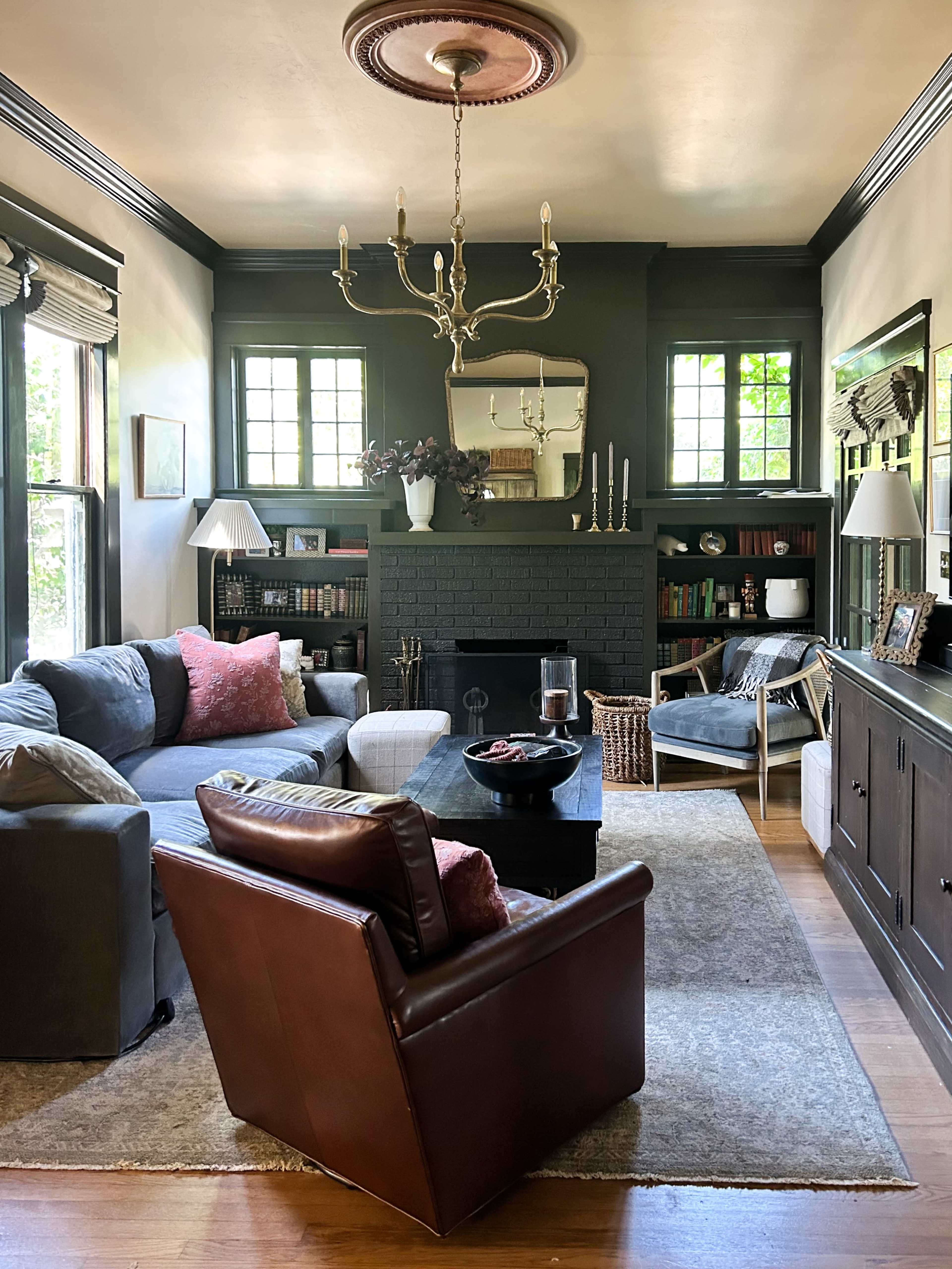The living room features a dark-colored fireplace, a large sofa with throw pillows, a leather armchair, and various decorative elements including a chandelier and bookshelves.