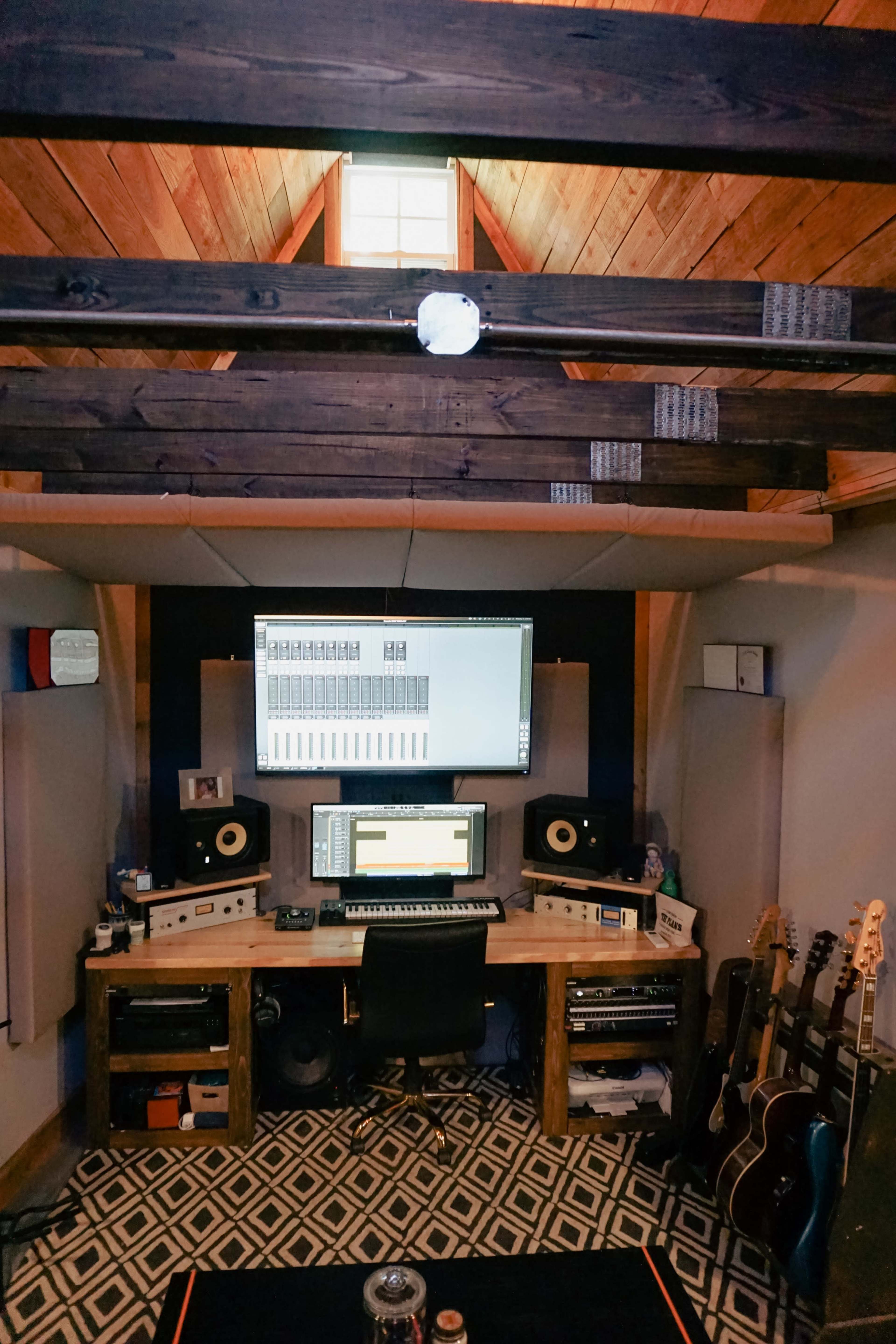 A music studio features a wooden desk with a computer monitor, audio equipment, and multiple guitars against the walls under a vaulted ceiling.
