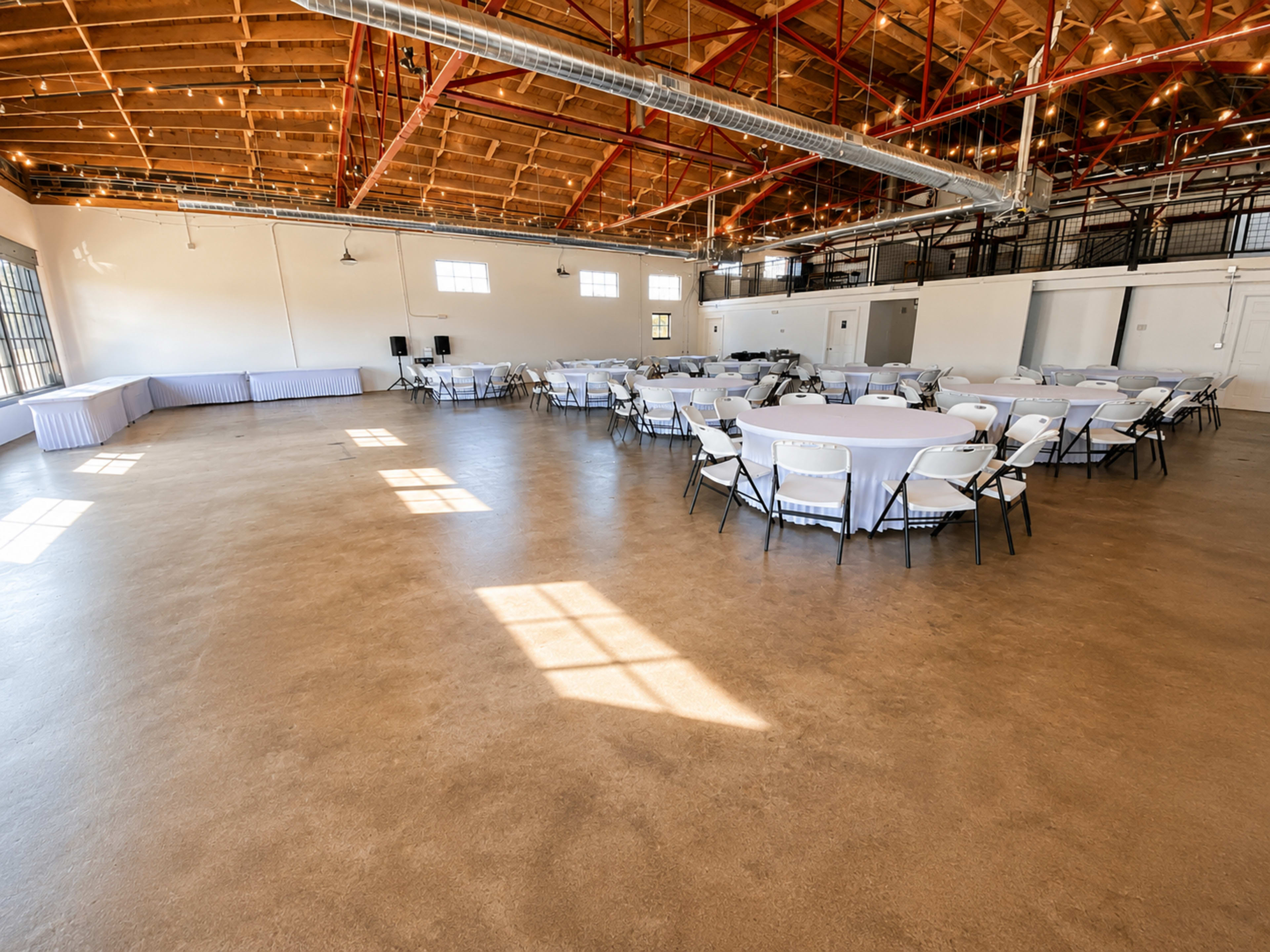 A spacious indoor event venue features several round tables set with white tablecloths, under a high ceiling with exposed wooden beams and a polished concrete floor.