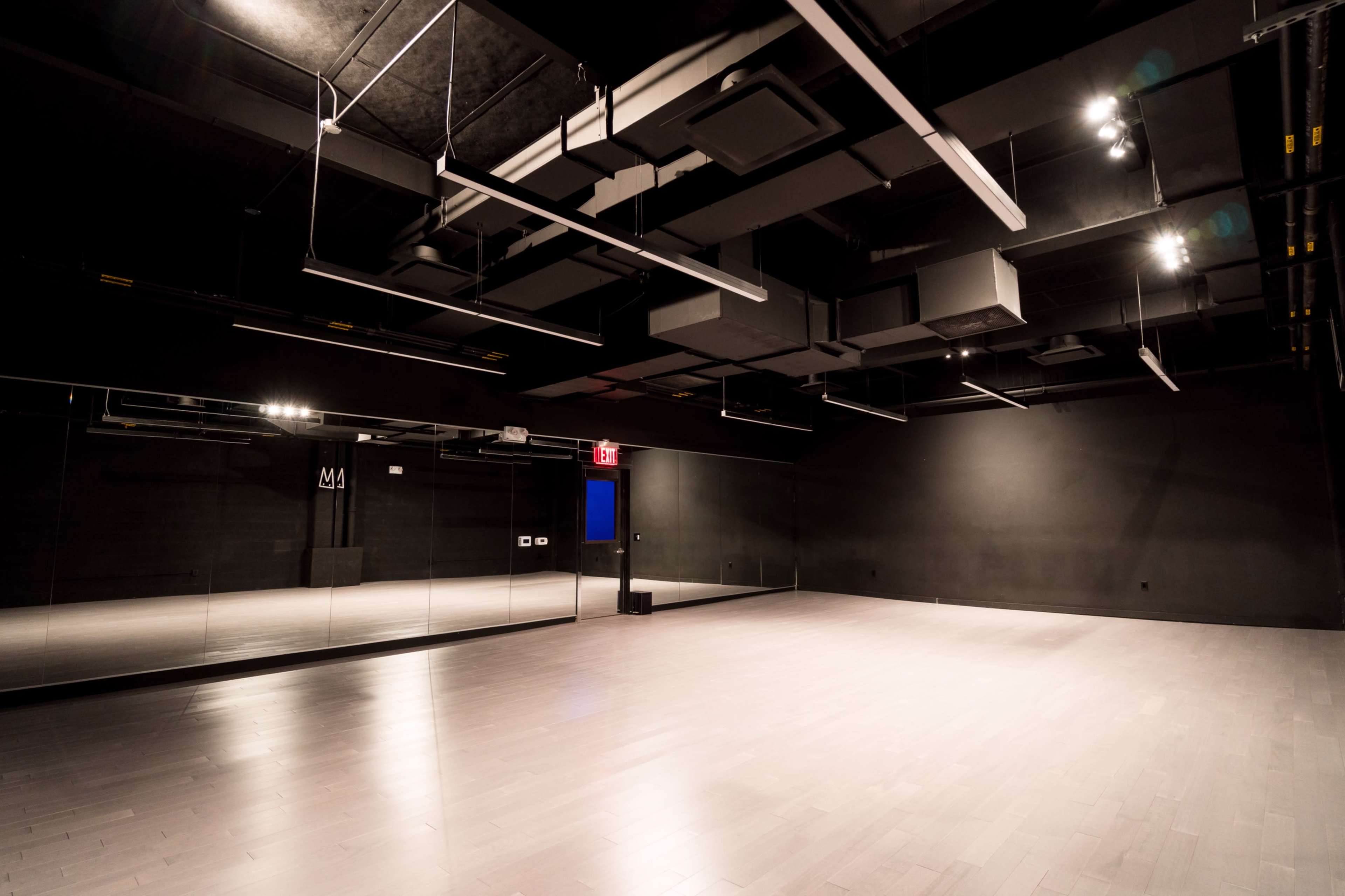 An empty dance studio featuring wooden flooring, black walls, and a large mirror on one side, with overhead lighting.