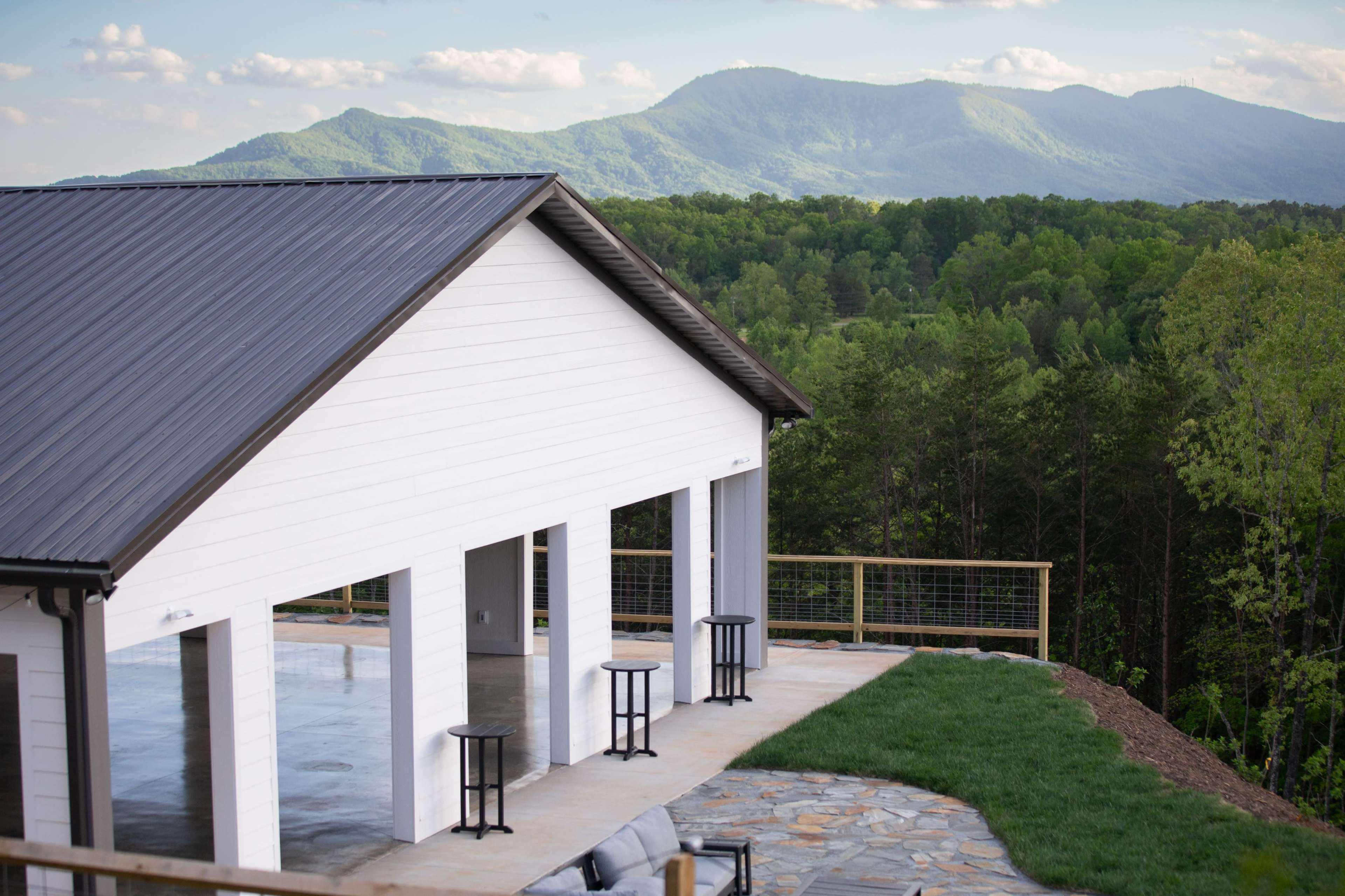 A modern white building with a metal roof overlooks a mountainous landscape, featuring expansive greenery and a stone patio with black stools.