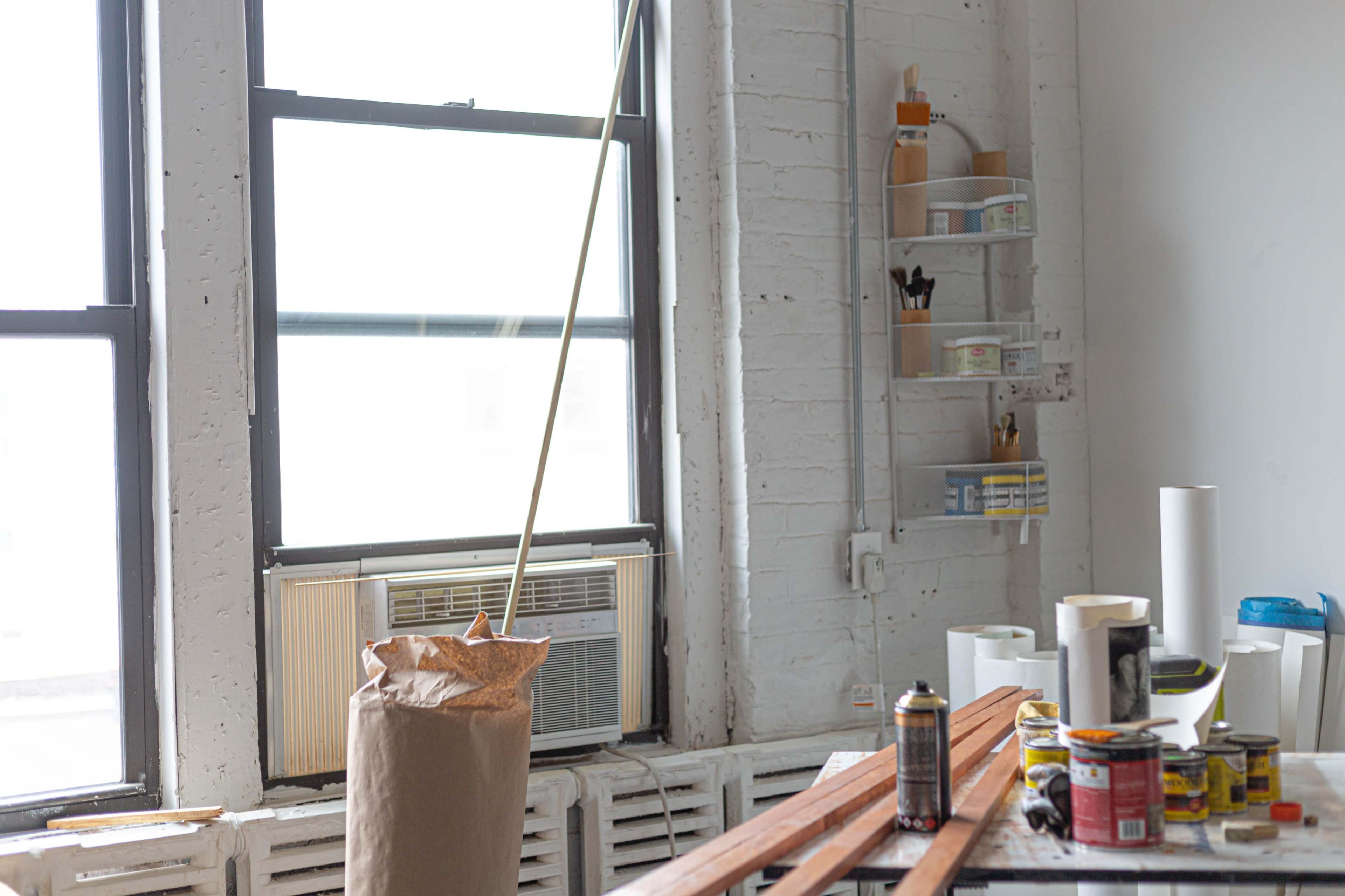 The image shows a bright room with a window, an air conditioning unit below it, and a cluttered table featuring various paint cans and supplies.