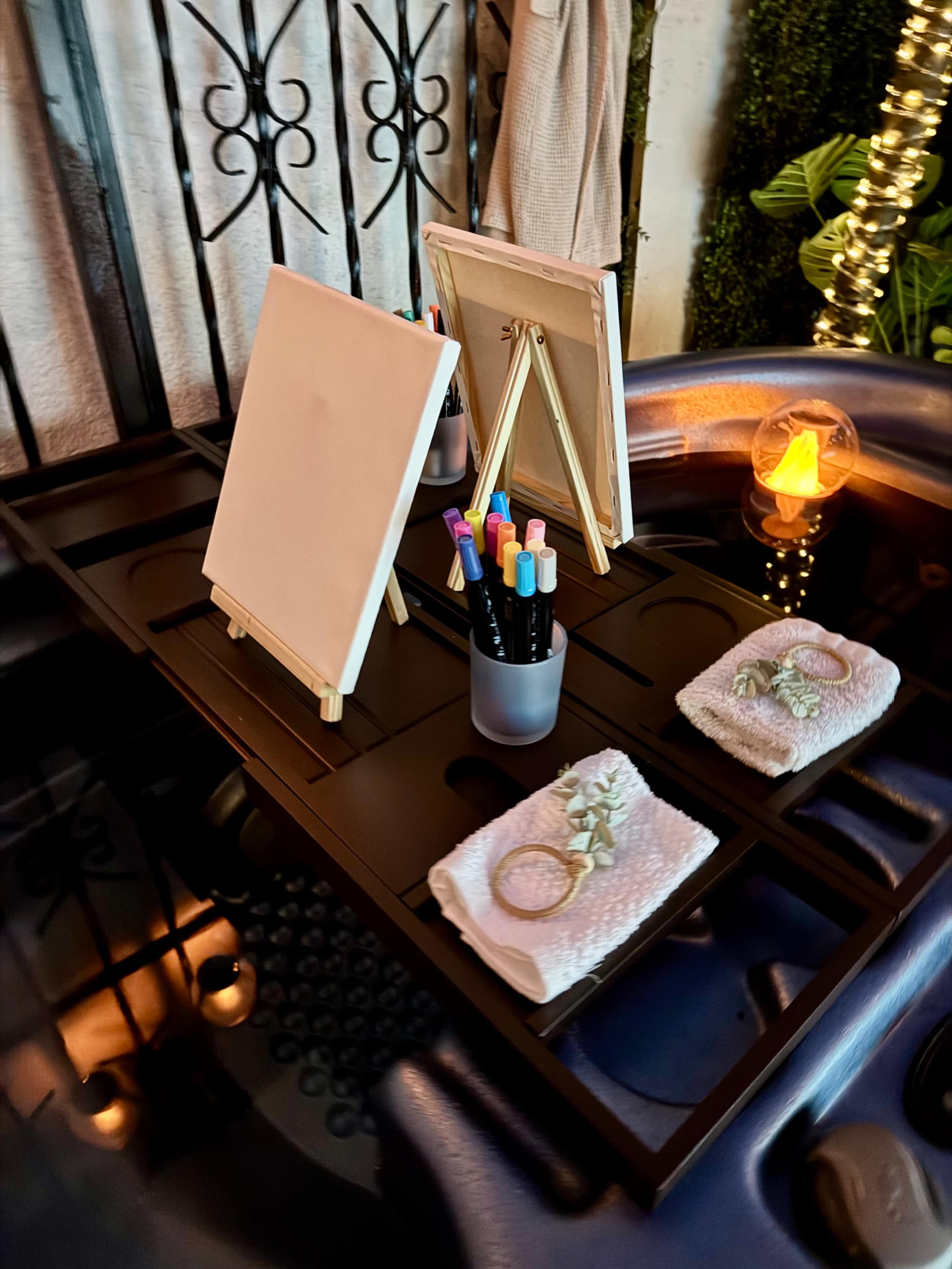 The image shows a small table with two blank canvases on easels, colorful markers in a cup, and neatly folded towels beside a glowing candle.