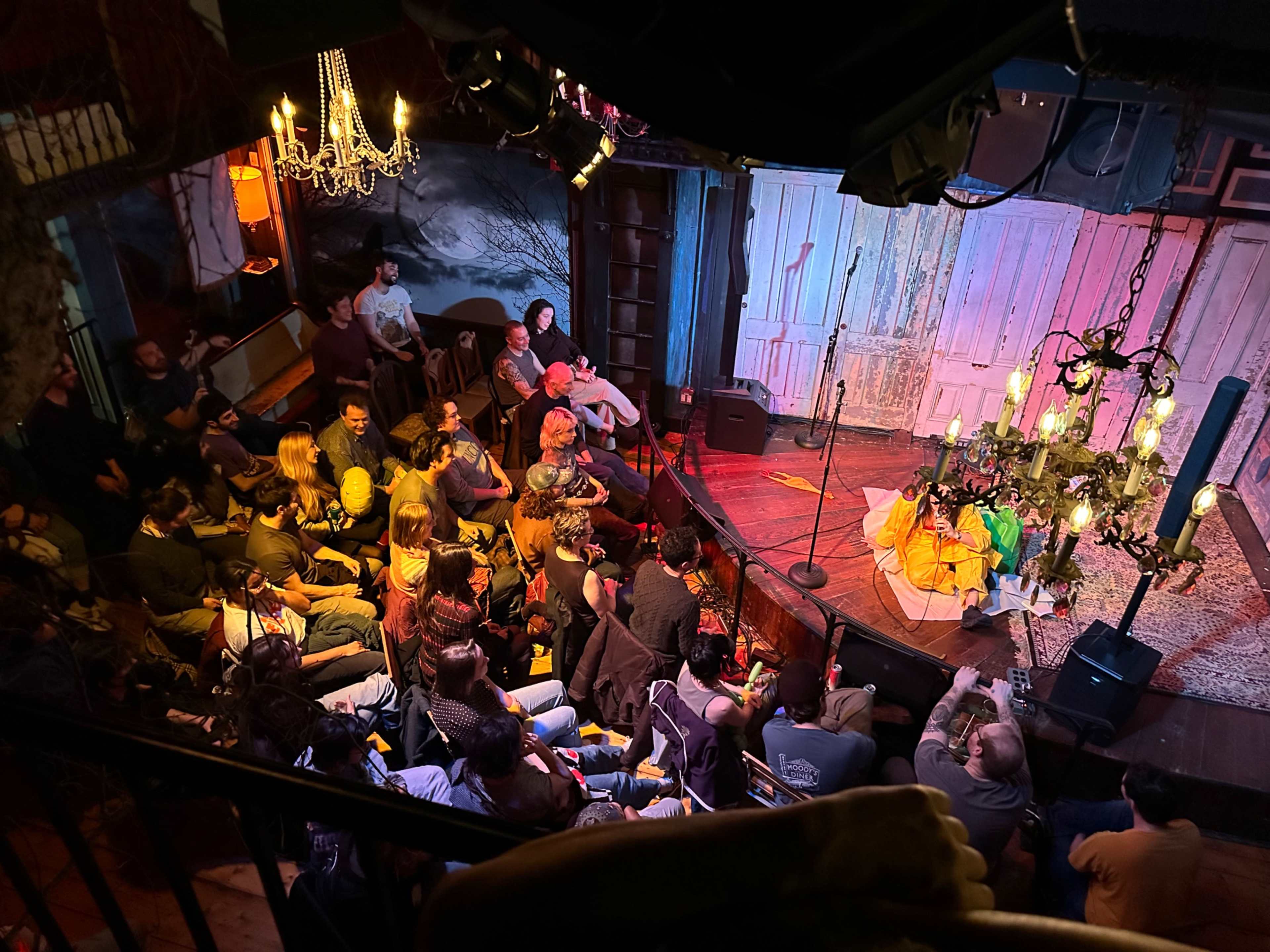 A crowd of people is seated in an intimate venue, facing a stage with a microphone and decorative lighting.