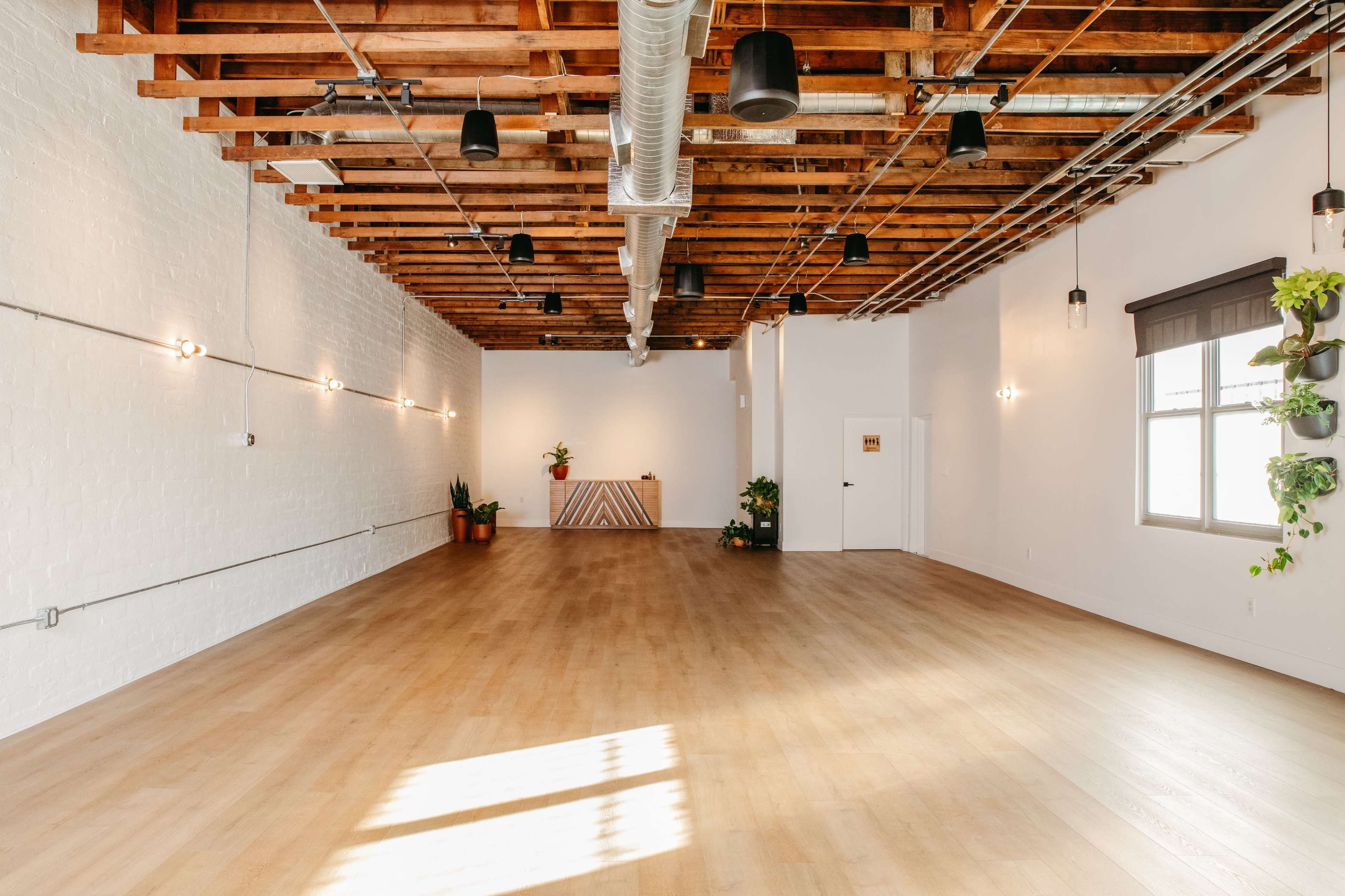 The image shows a spacious, empty room with wooden floors, exposed beams, and minimal decor, featuring plants and light fixtures on the walls.