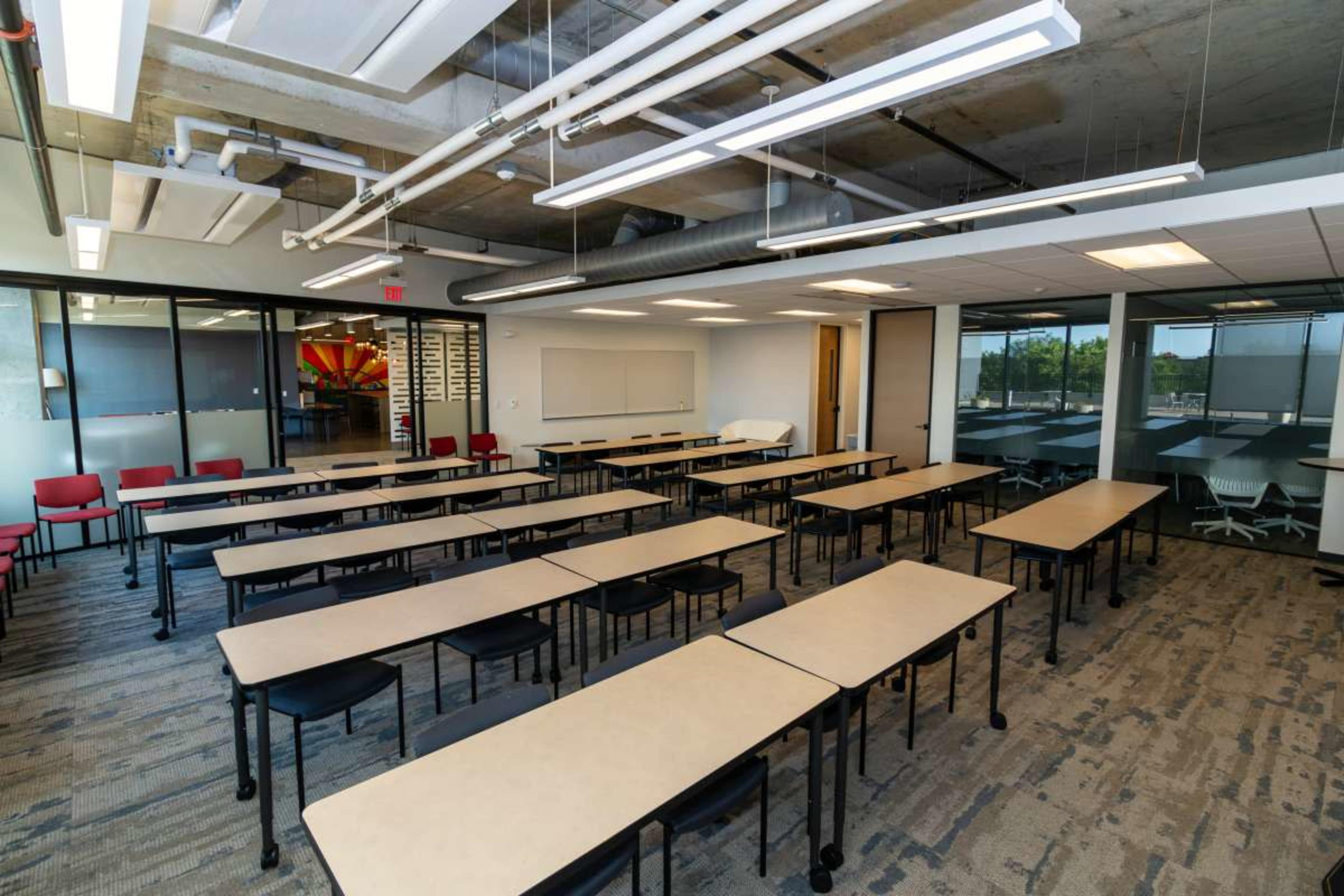 The image shows a modern classroom with rows of tables and chairs arranged for use, featuring large windows and overhead lighting.