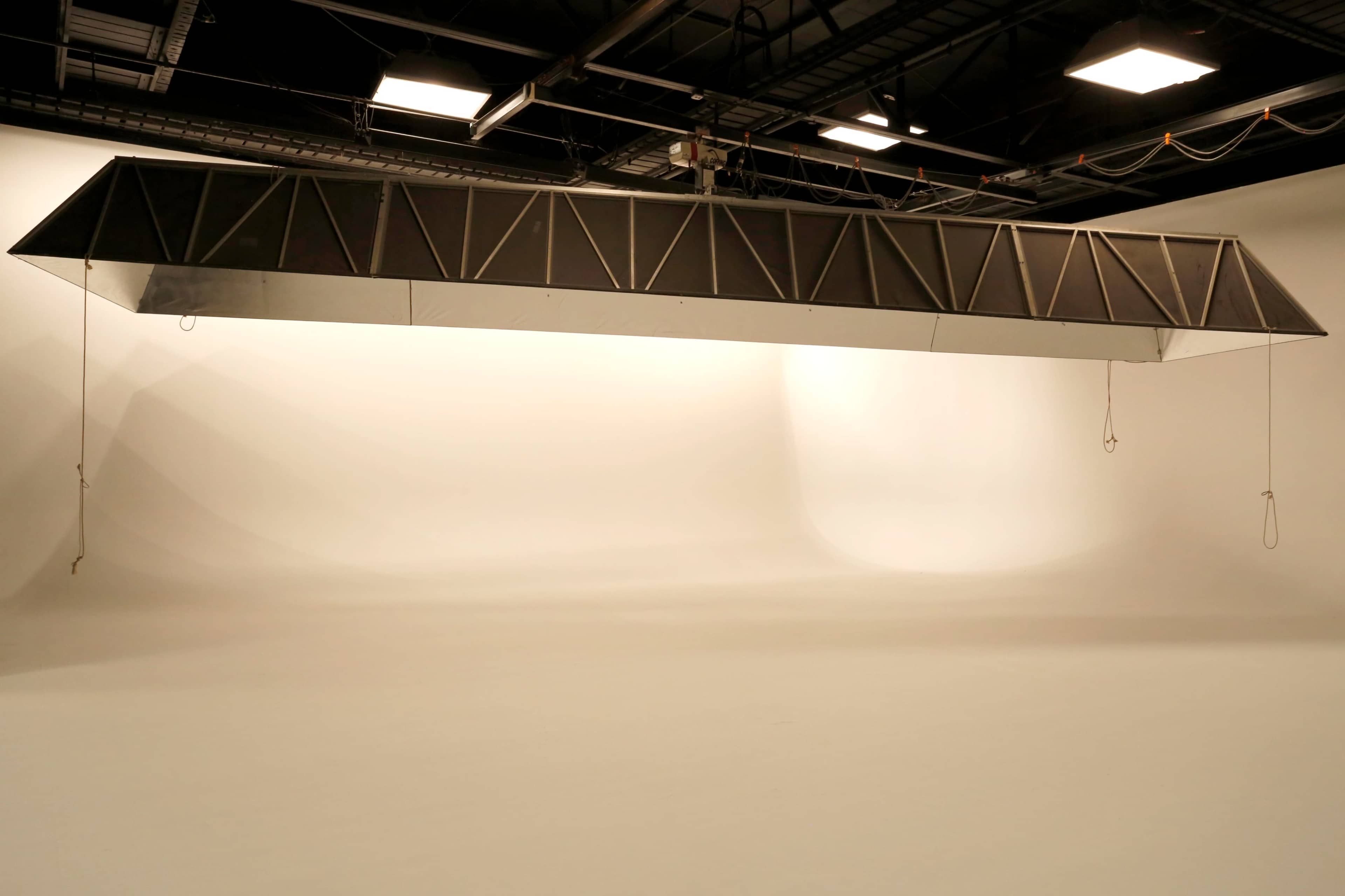 A large, suspended lighting rig hangs from the ceiling in a spacious photography studio with a plain white backdrop.