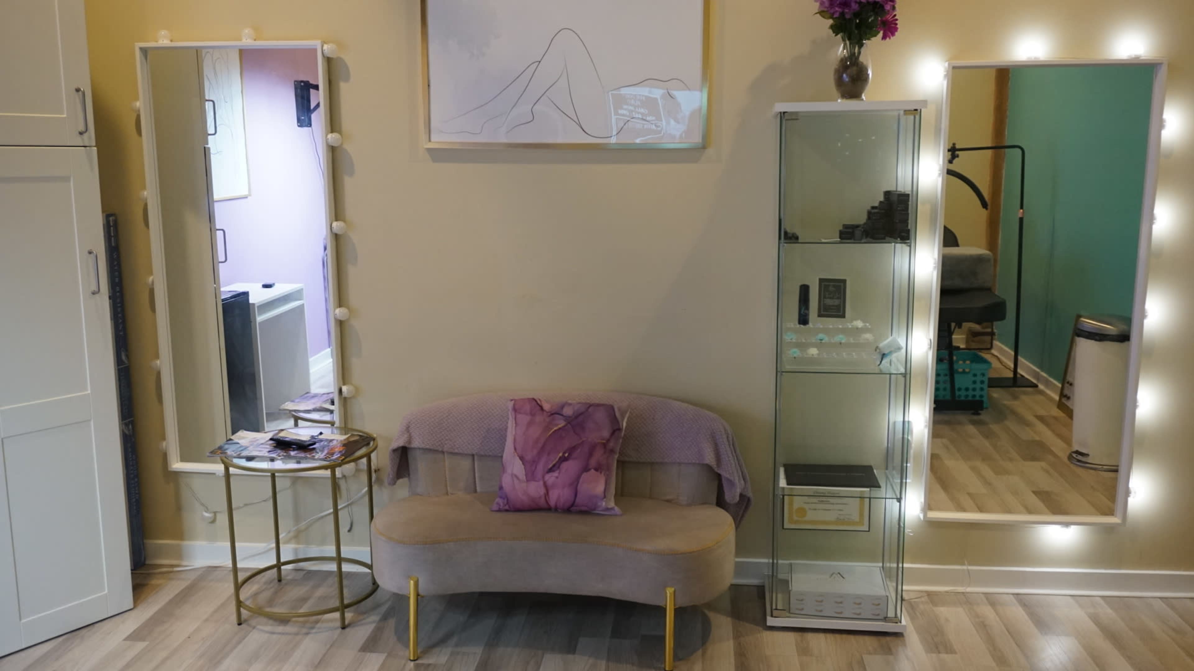 The image shows a cozy seating area in a salon, featuring a curved bench with a purple pillow, two mirrors, and a glass display case with various items inside.