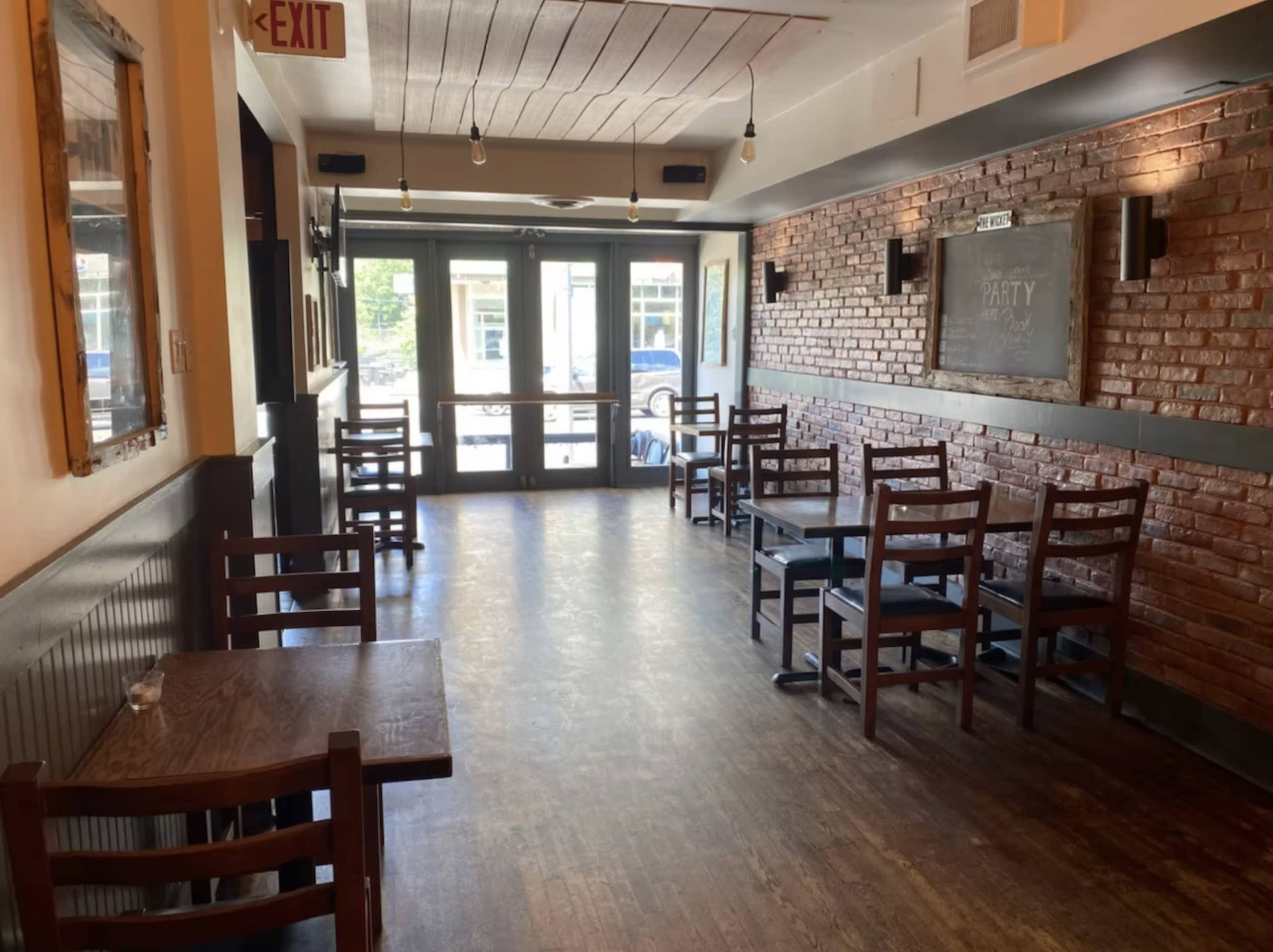 The image shows an empty restaurant interior featuring wooden tables and chairs, with exposed brick walls and large glass doors letting in natural light.