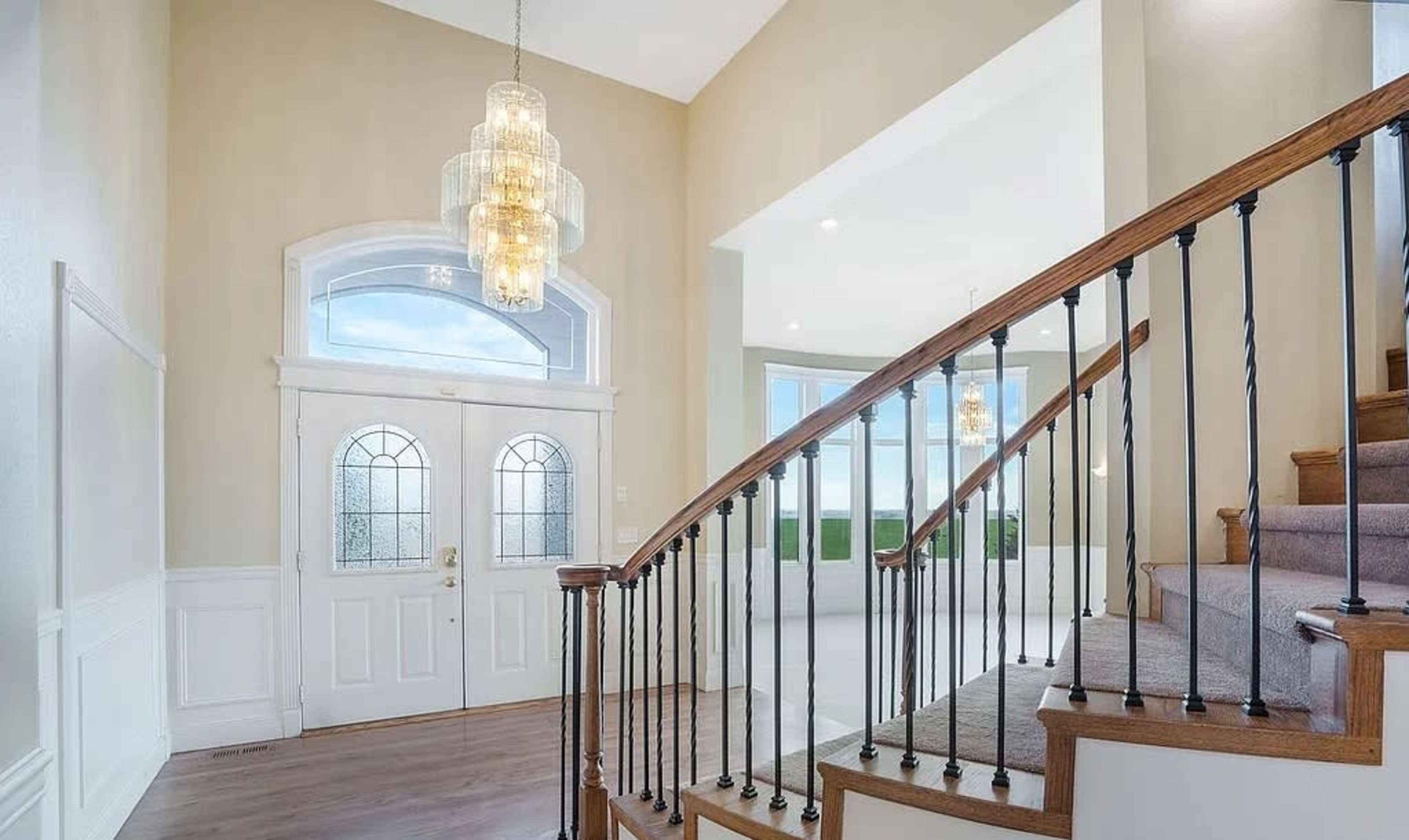 The image shows a wooden staircase with a wrought iron railing leading to a bright, open foyer featuring large windows and a chandelier.