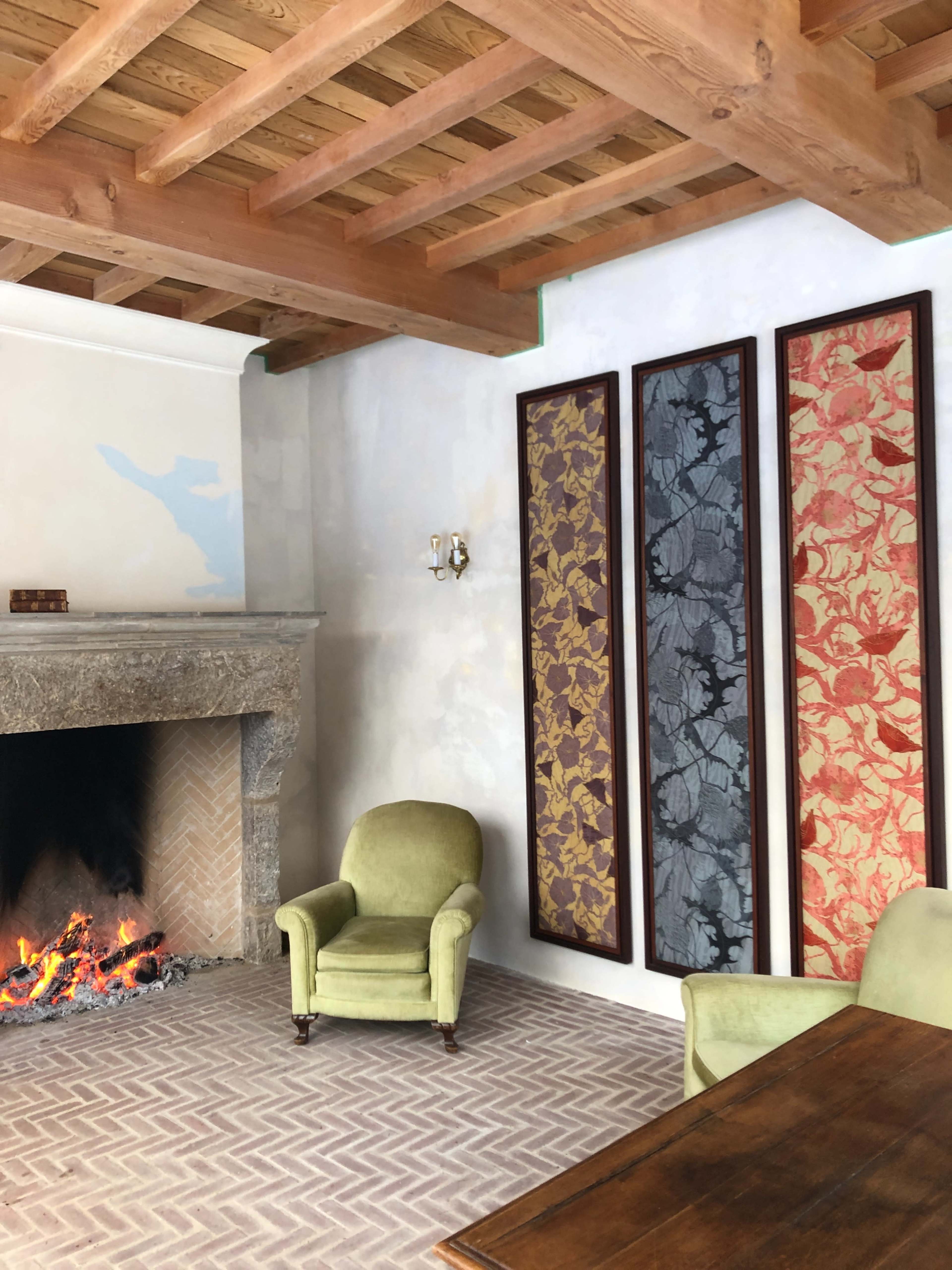 A cozy room featuring a stone fireplace with a lit fire, two green armchairs, a wooden table, and three decorative framed panels on the wall.