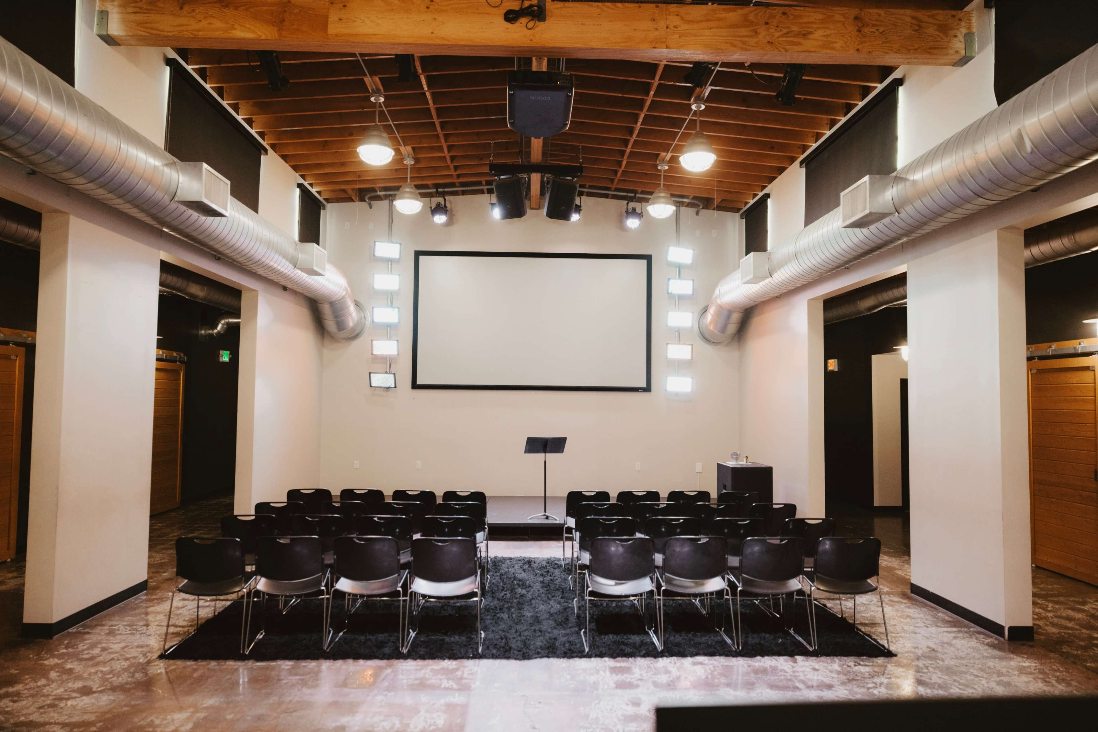 A modern presentation room features rows of black chairs facing a large screen, with a podium at the front and overhead lights along the walls.