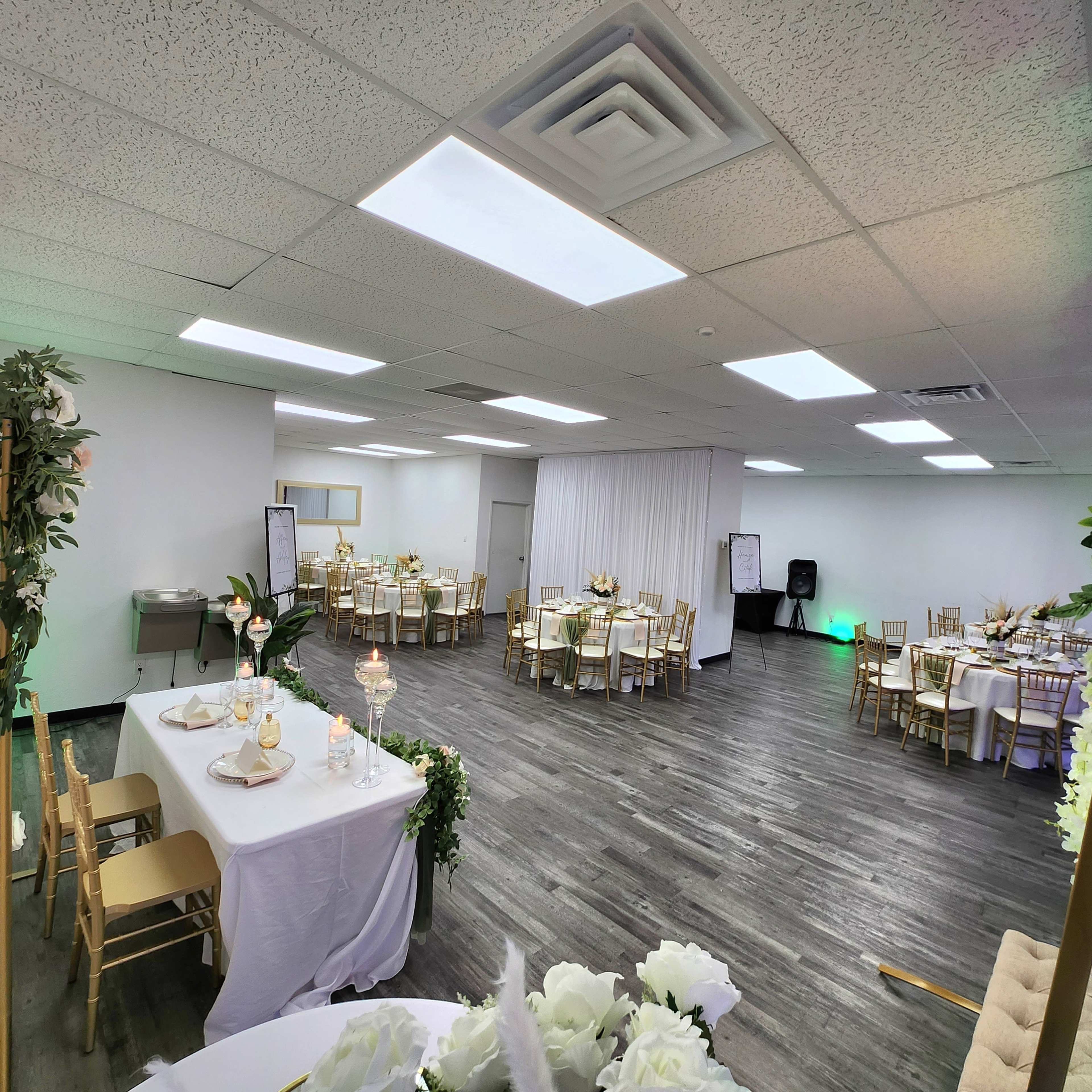 A spacious event room arranged with tables and chairs set for a gathering, featuring white tablecloths and floral centerpieces.