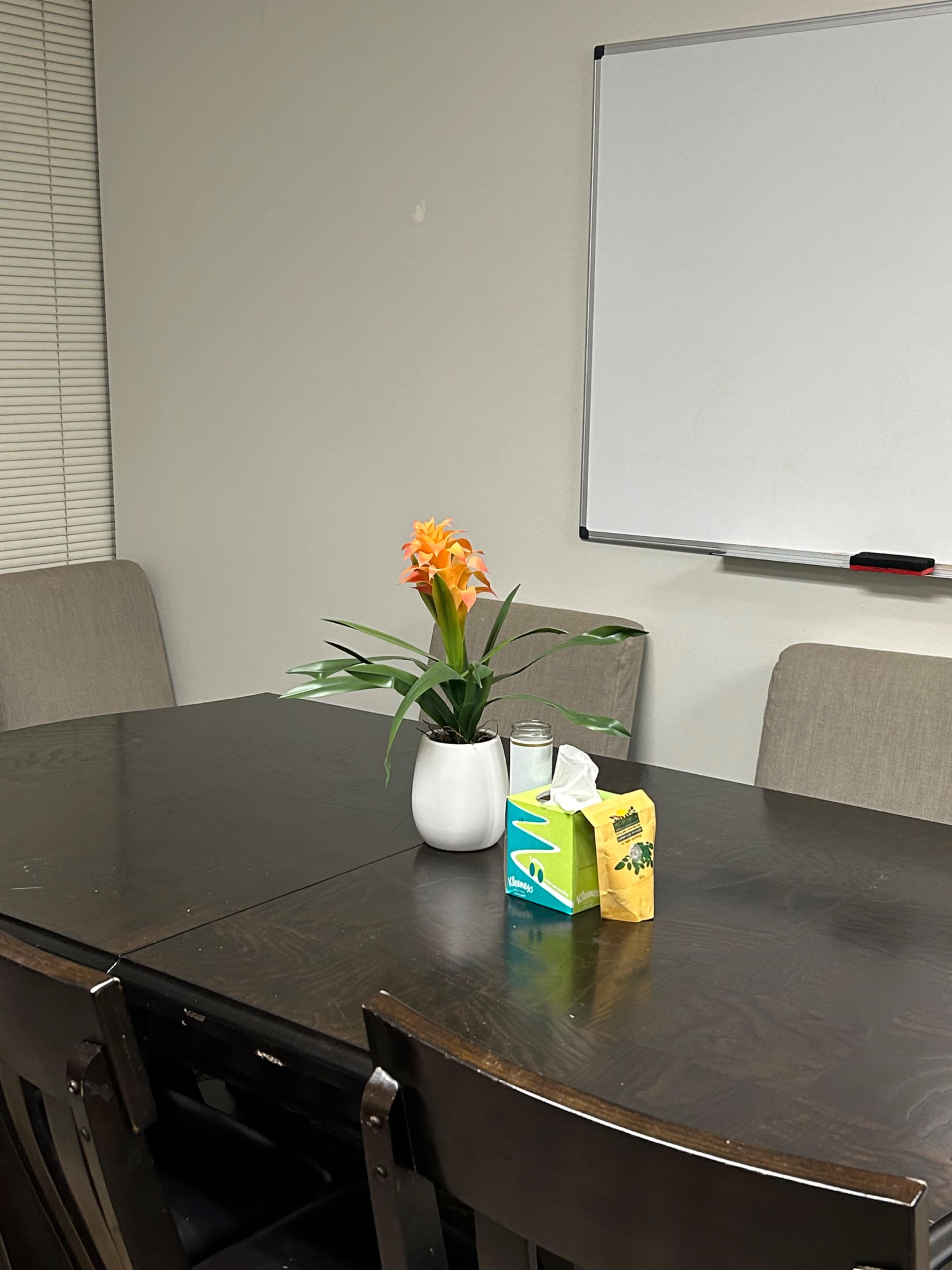 A small conference table is set with a potted flower, a box of tissues, and a carton of juice, with gray chairs positioned around it and a whiteboard in the background.
