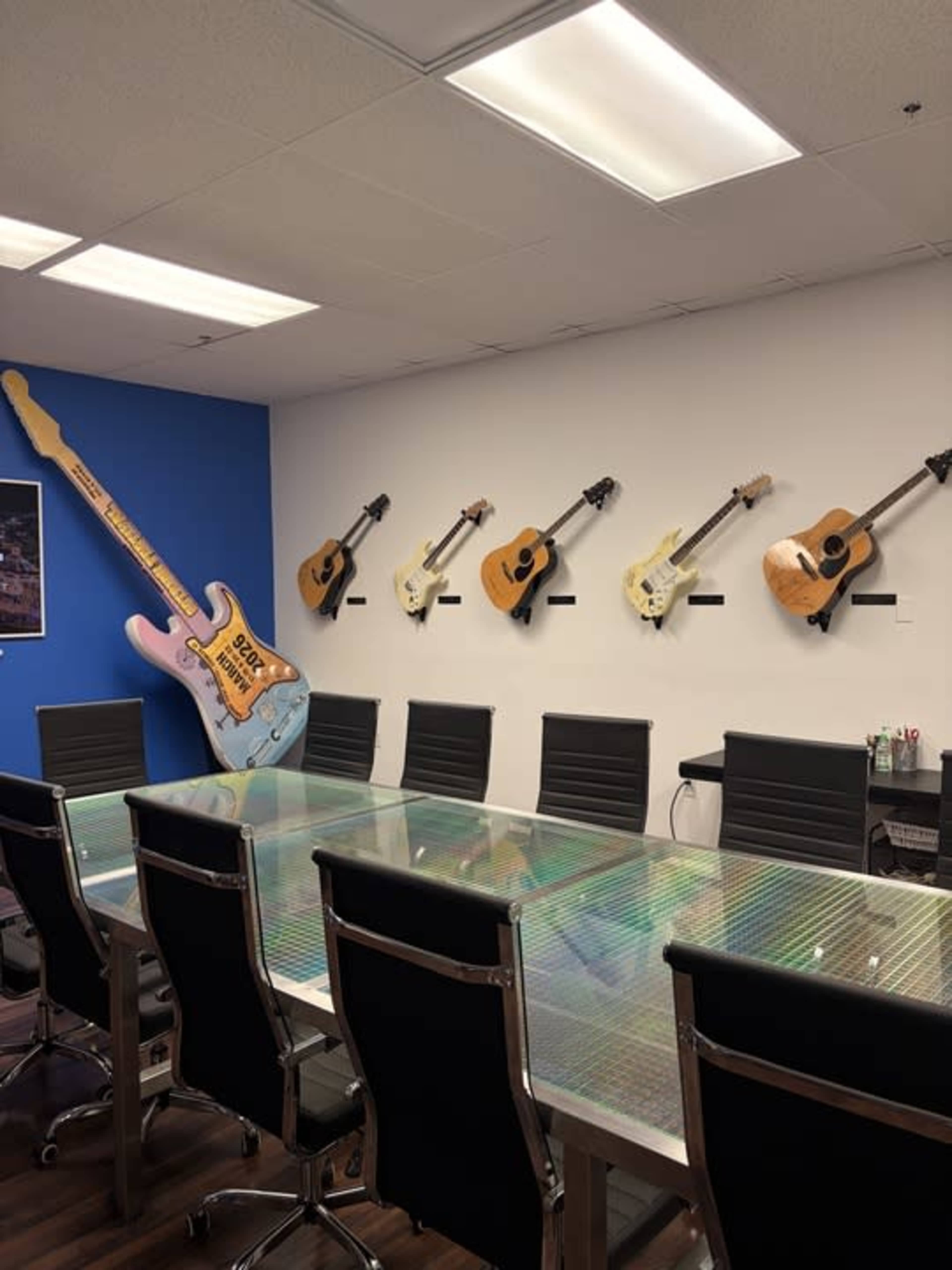 The image shows a conference room with a large glass table and black chairs, featuring several guitars mounted on the walls, including an oversized pink guitar on the left.