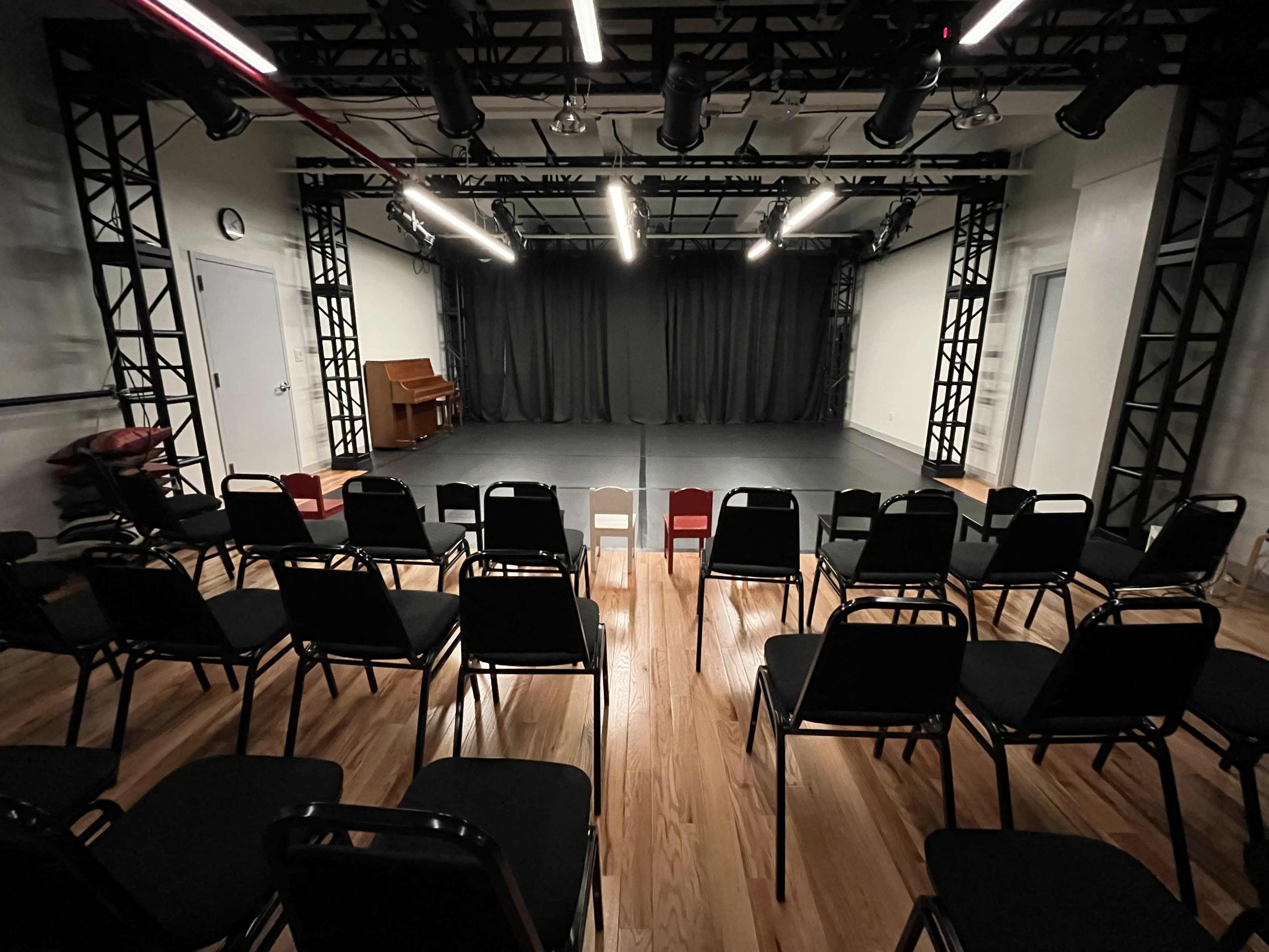 A theater rehearsal space with a seating area of black chairs facing a black curtain and a wooden piano on the side.