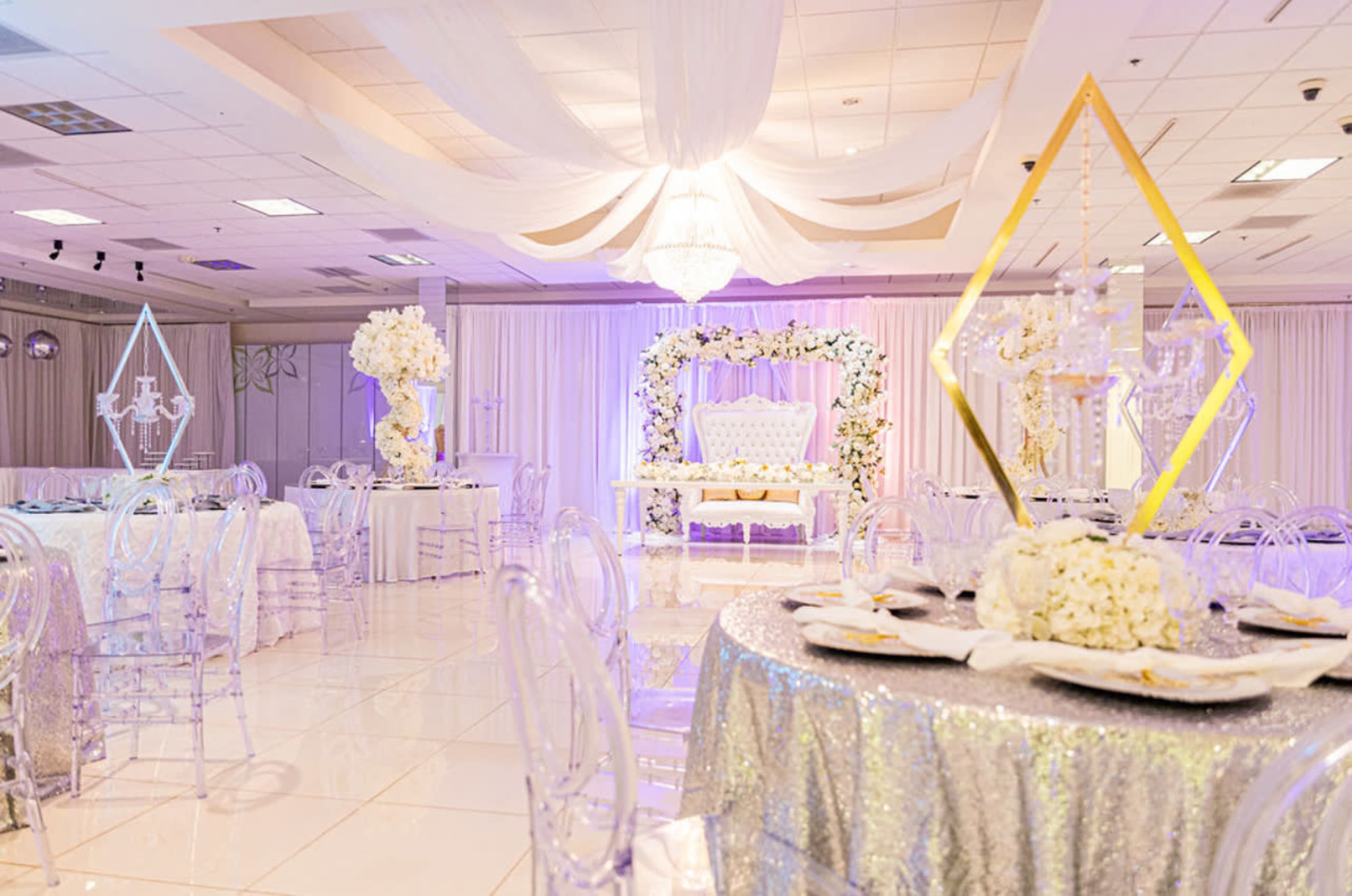 The scene shows a banquet hall decorated with elegant tables, transparent chairs, and floral arrangements, featuring a large backdrop with a throne and draping overhead.
