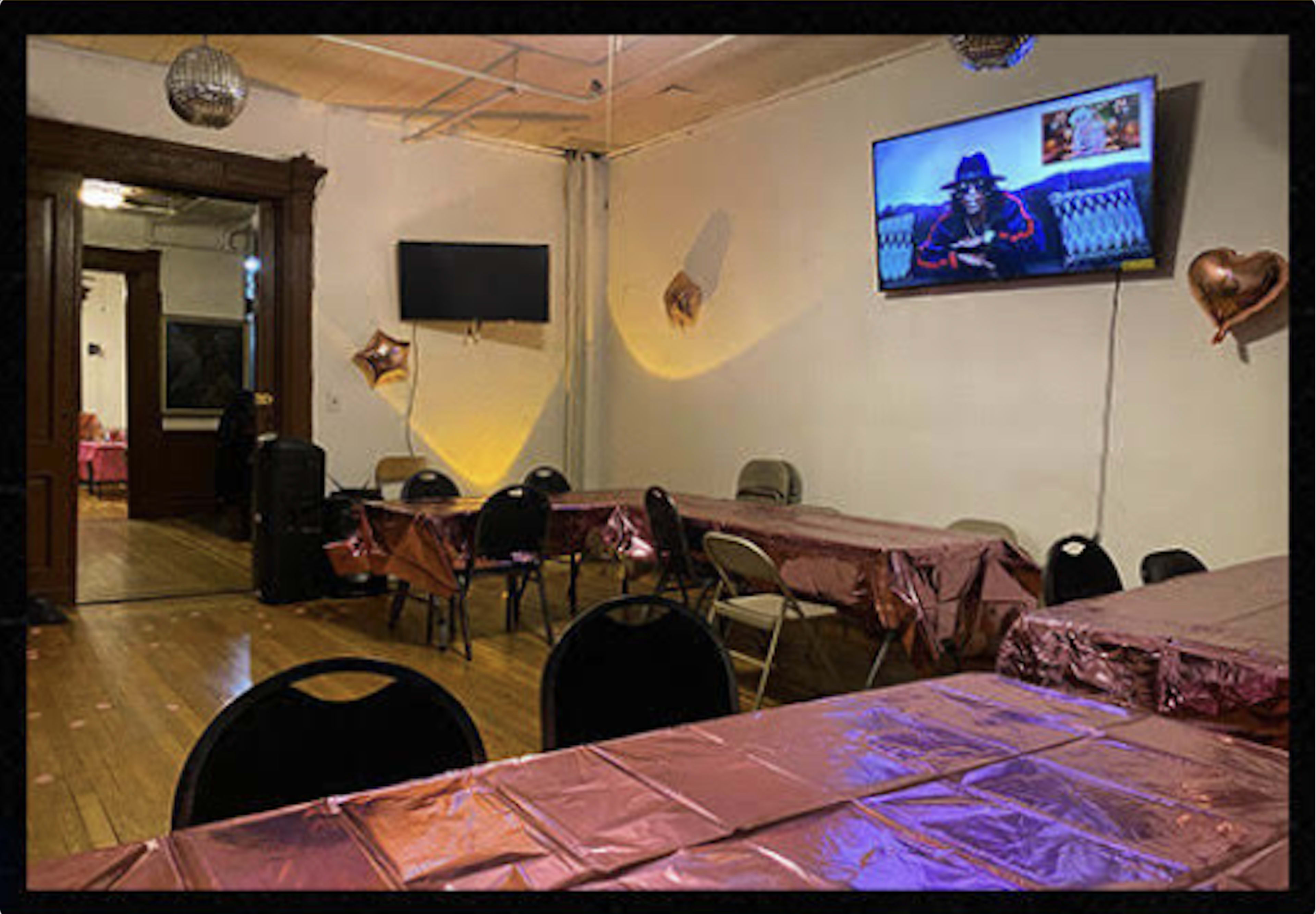 The image shows a plain event room with tables covered in pink tablecloths, decorated with heart-shaped balloons, and a television displaying an image on the wall.